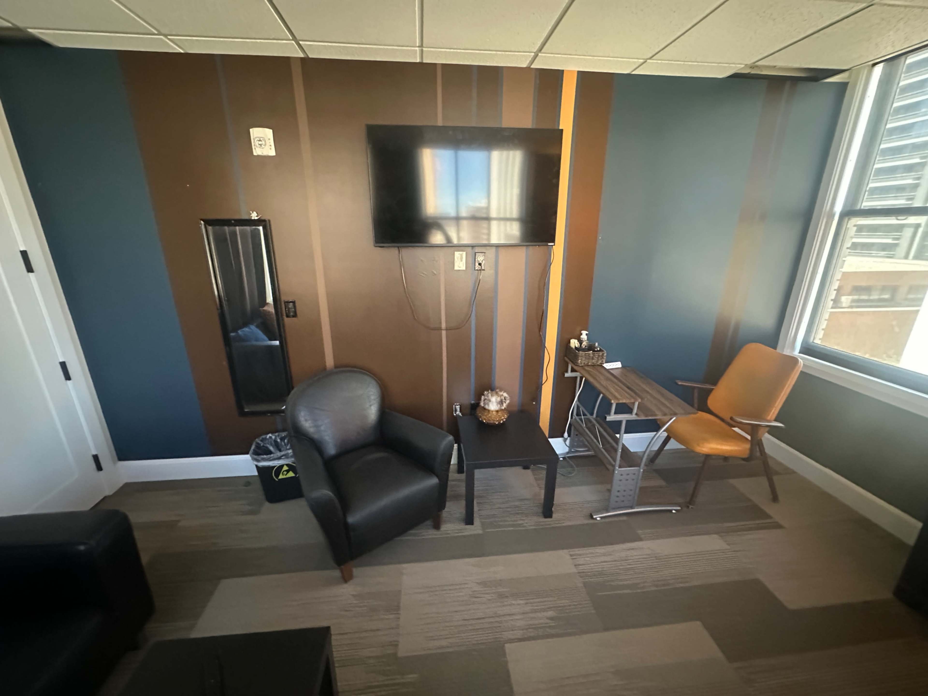 The image shows a small office or waiting area with a television mounted on the wall, a black chair, a wooden table, and an orange chair.