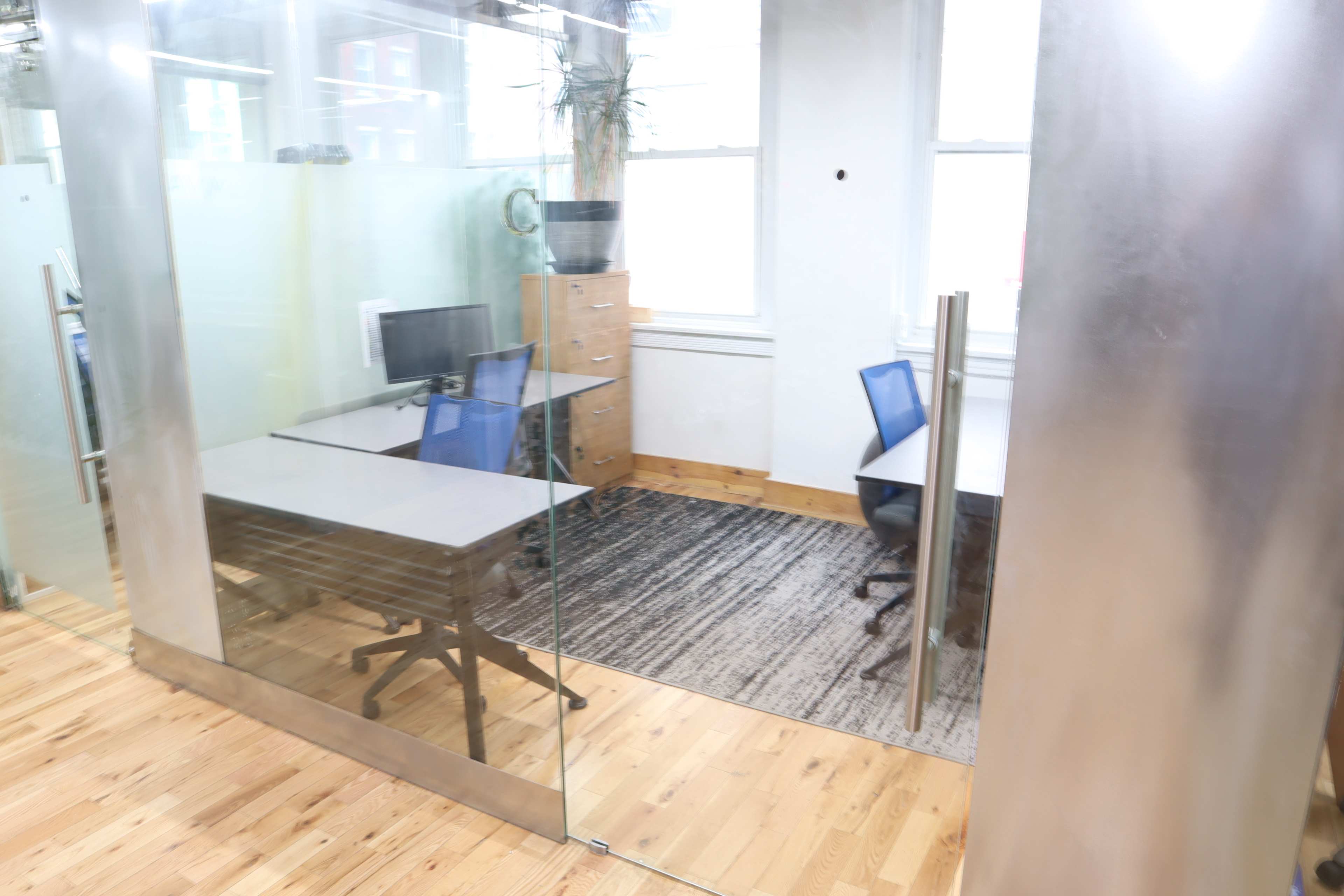 The image shows a modern office space with two desks and chairs separated by glass partitions.
