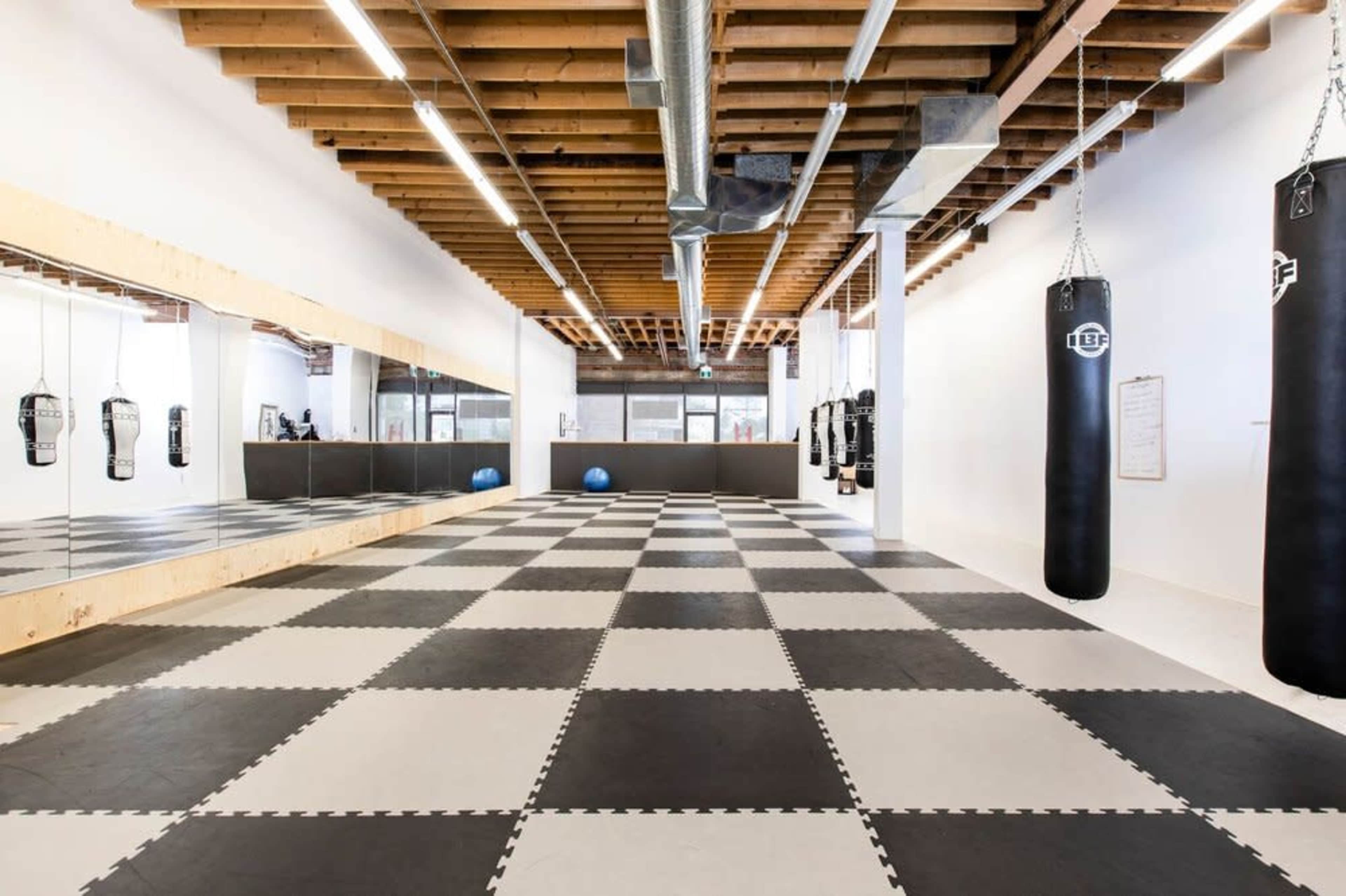 A spacious, empty training area with black and white checkered flooring, reflective mirrors on one wall, and several hanging punching bags.