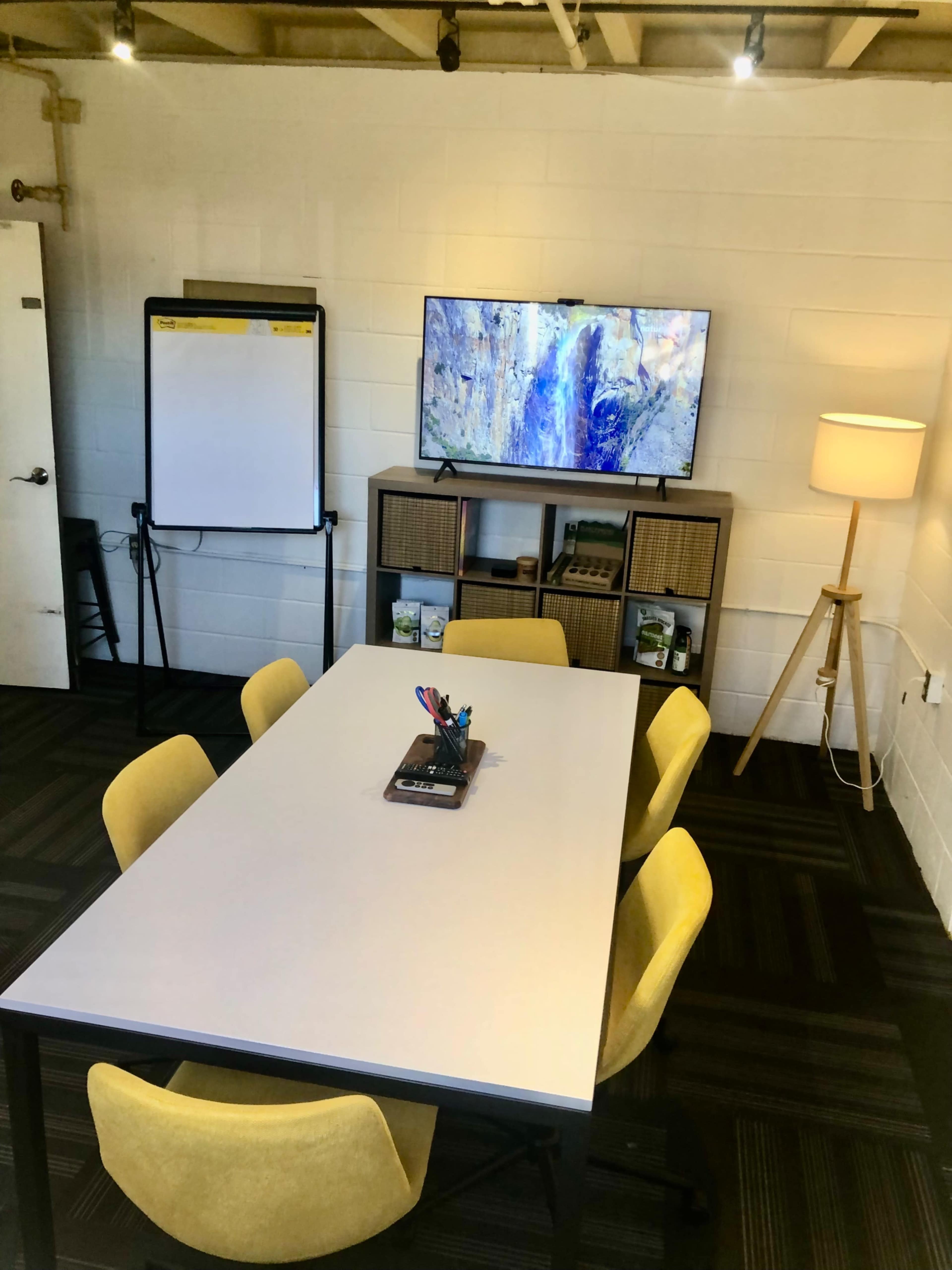 A meeting room features a white table surrounded by yellow chairs, a TV mounted on the wall, a flip chart, and a lamp in the corner.