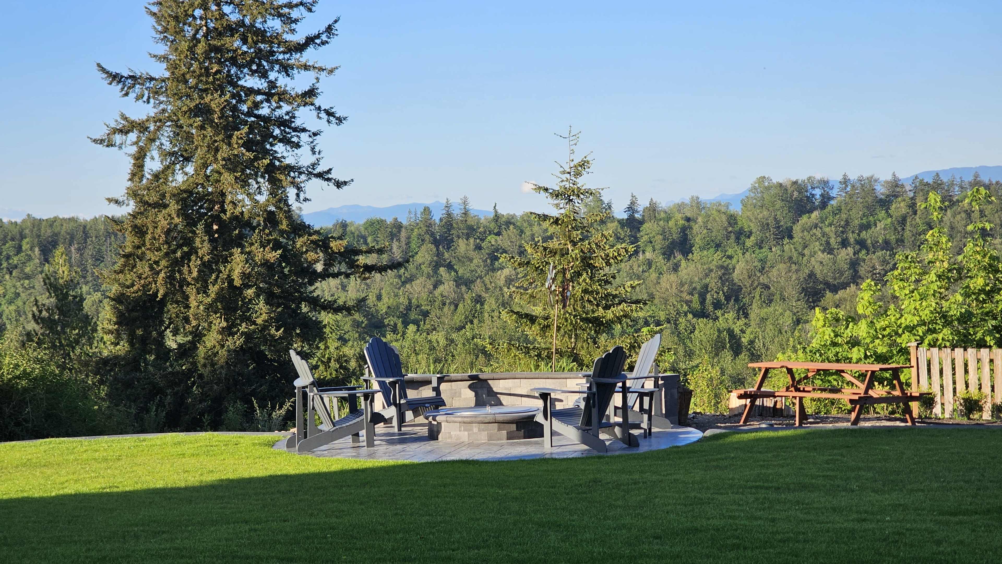 A circular stone fire pit is surrounded by wooden chairs on a grassy area, with a wooded landscape and mountains in the background.