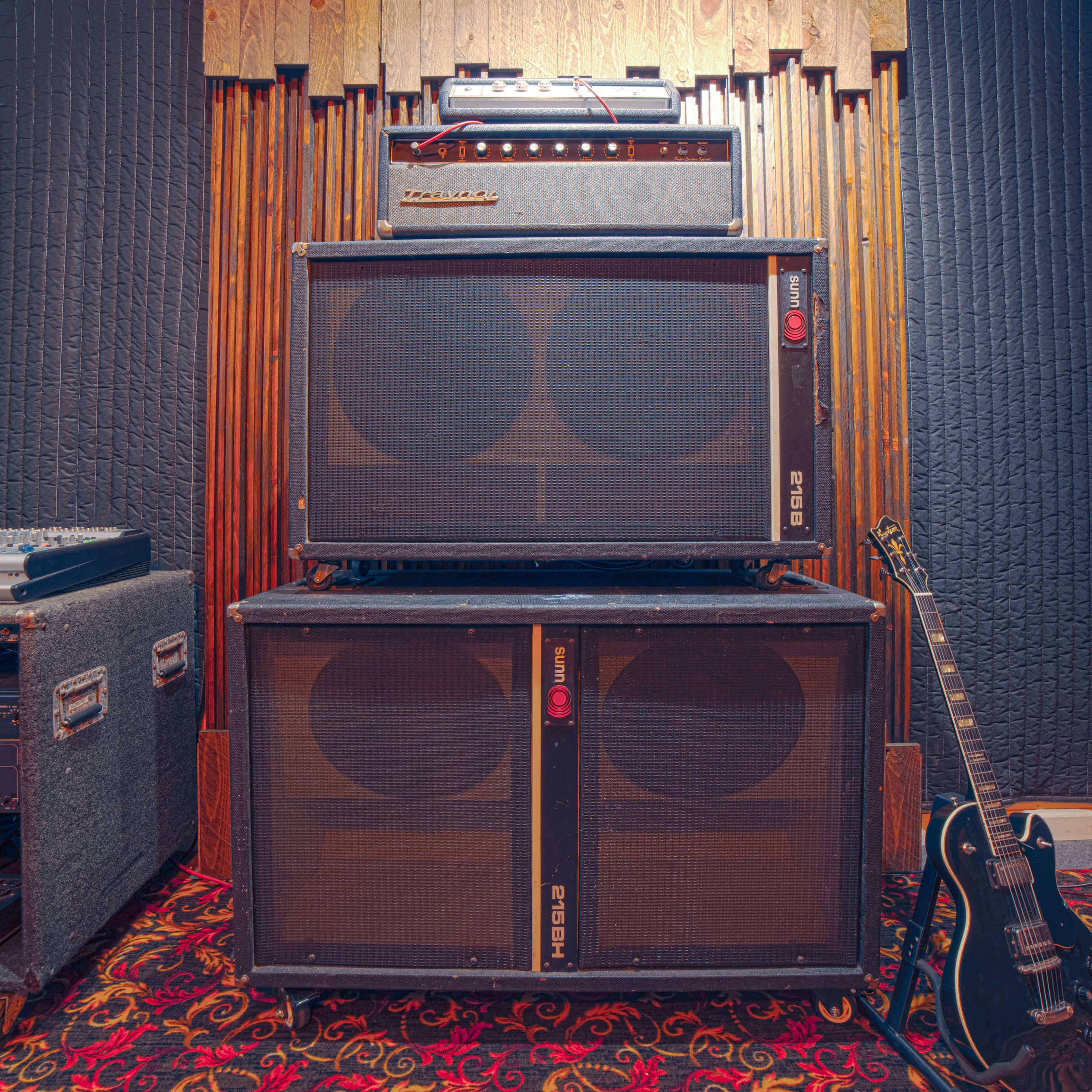 The image shows a stack of amplifiers and speaker cabinets next to an electric guitar in a recording studio setting.