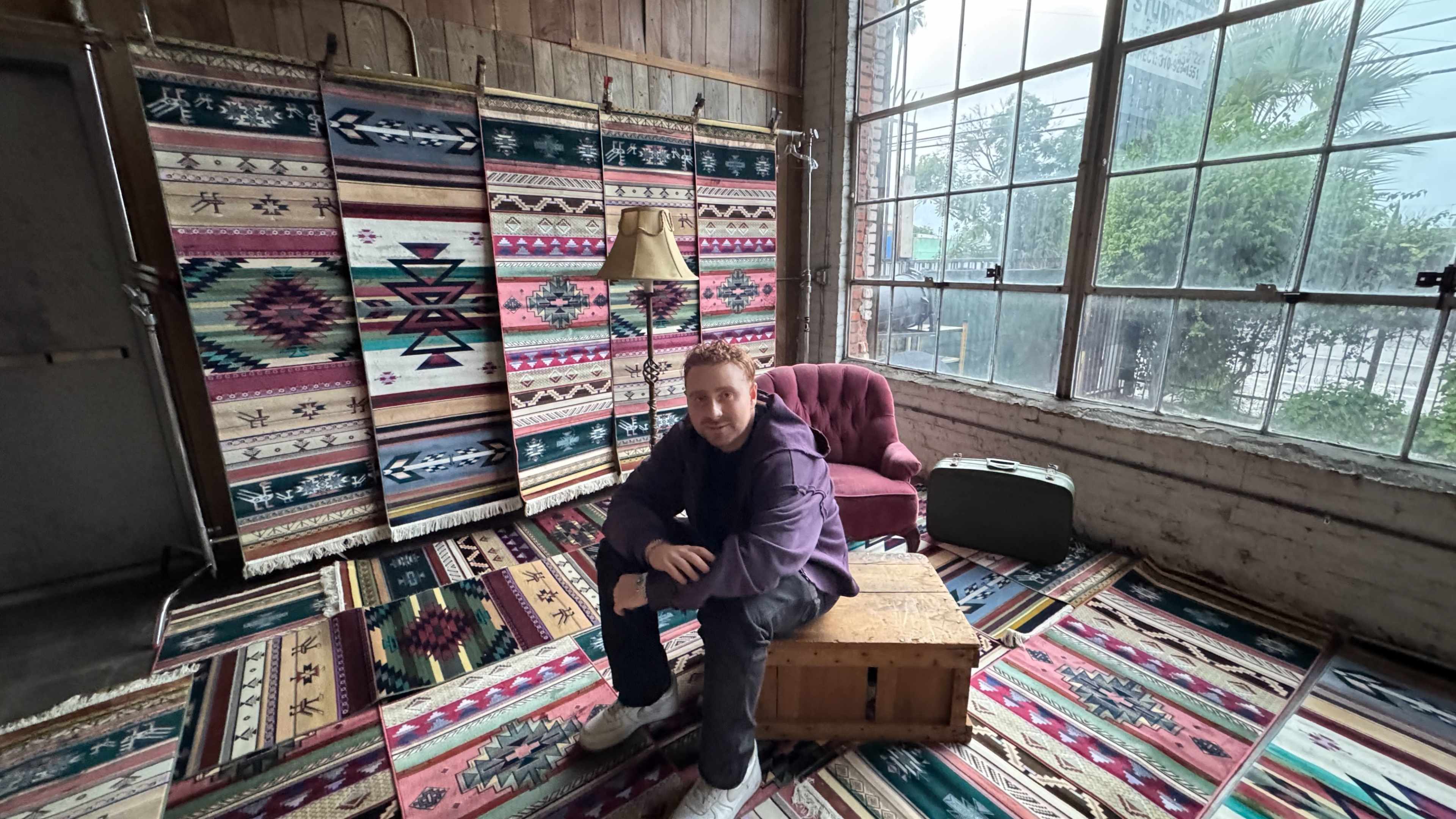 A person sits on a wooden crate in a room with vibrant patterned rugs and wall hangings, accompanied by a lamp and a burgundy couch.