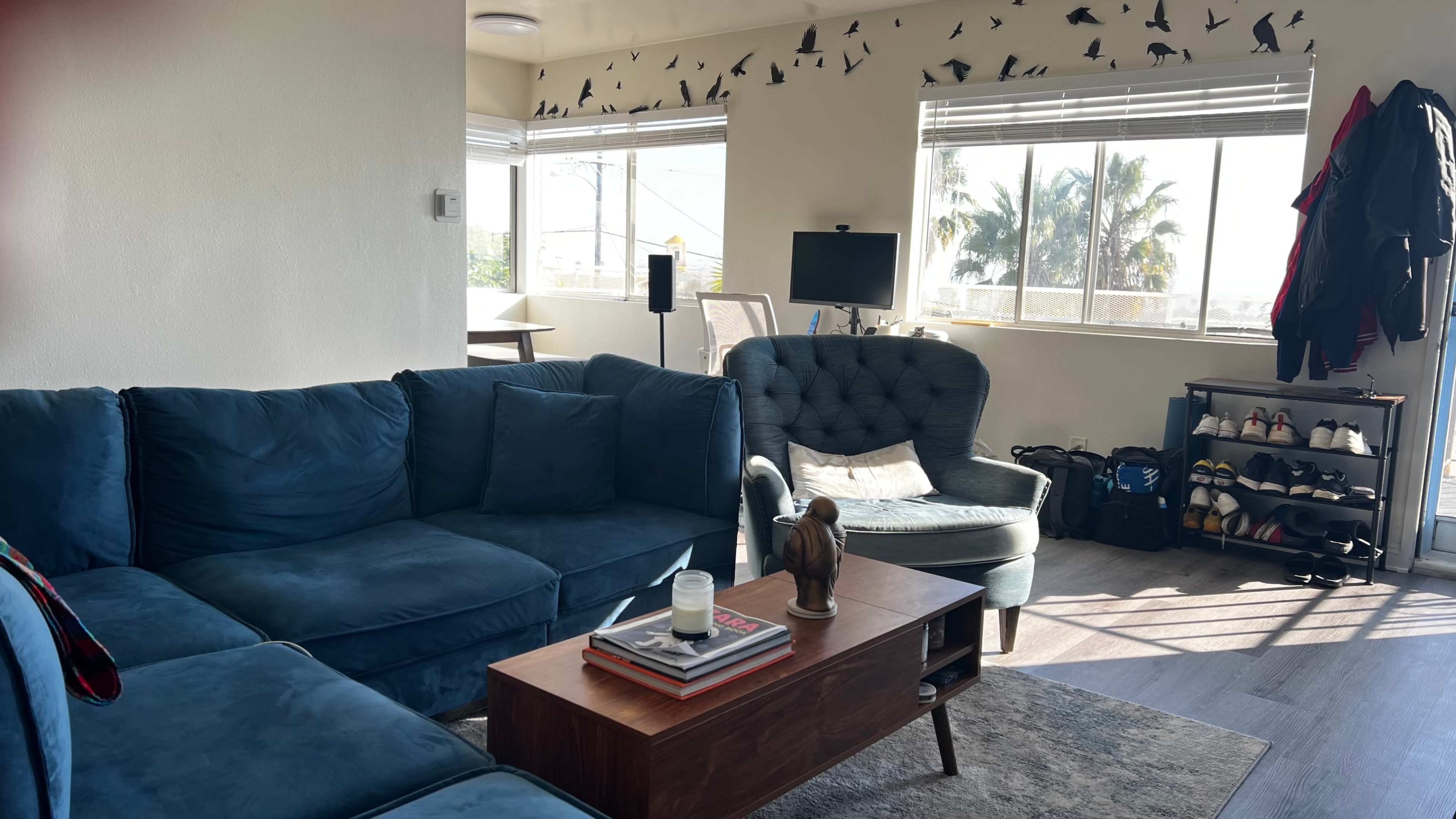The image shows a living room with a blue sectional couch, a coffee table with books and a candle, a green armchair, and a small table with a computer in front of a window.