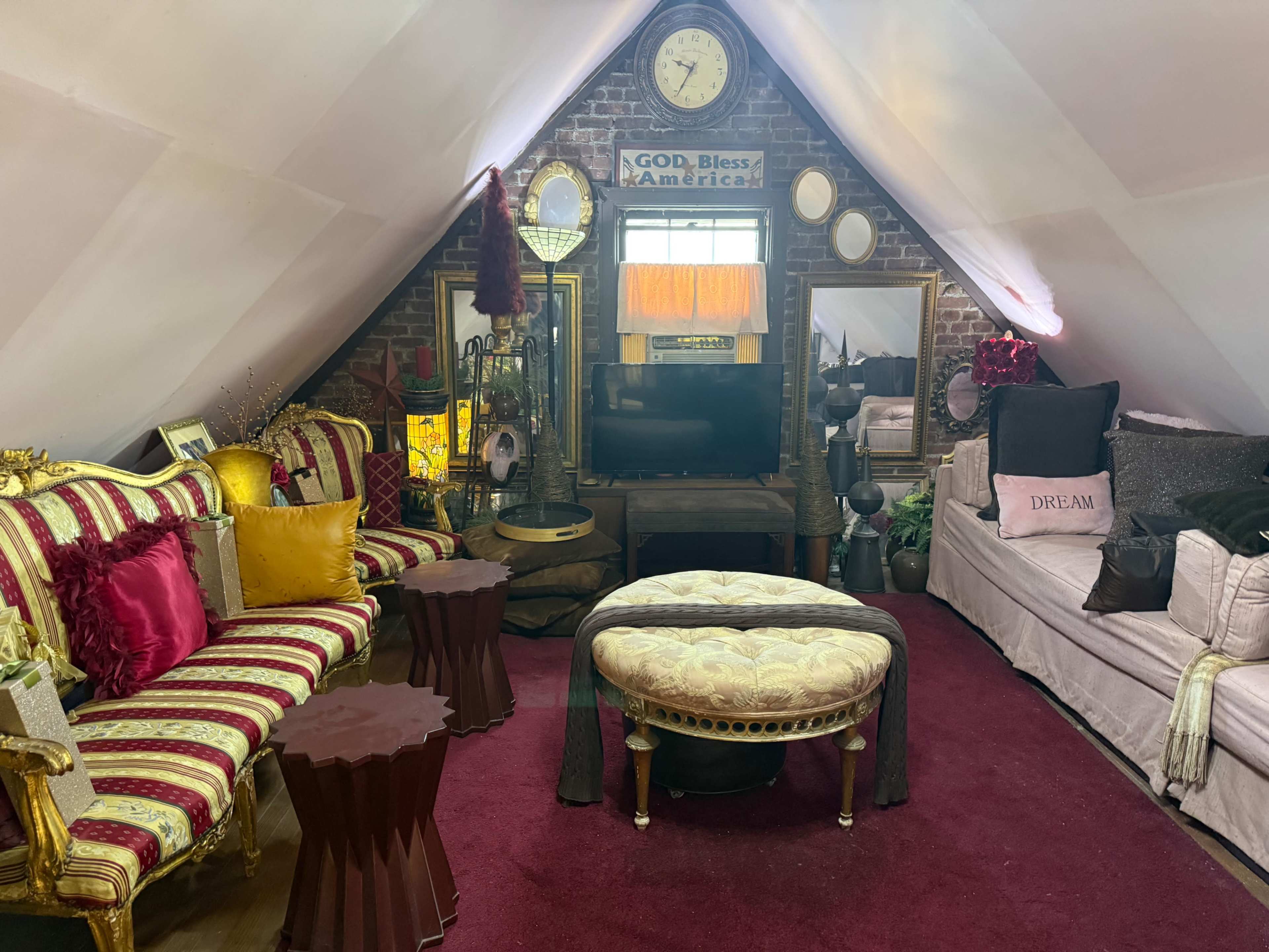 The image shows a cozy attic space furnished with vintage-style sofas, a round ottoman, and decorative accents, including a clock and various plants.