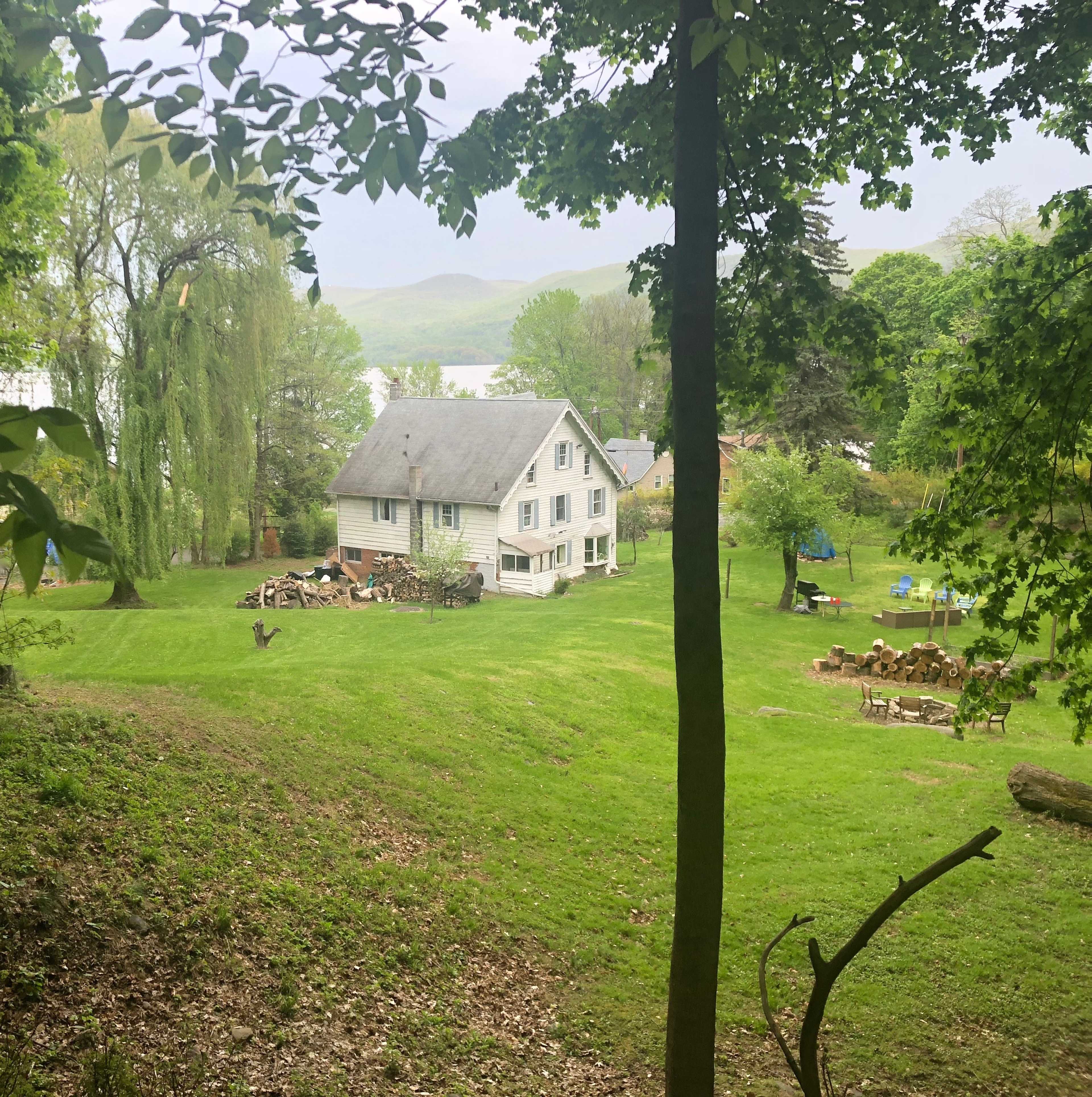 A two-story white house is situated on a grassy area surrounded by trees, with a yard that includes stacked firewood and outdoor seating.