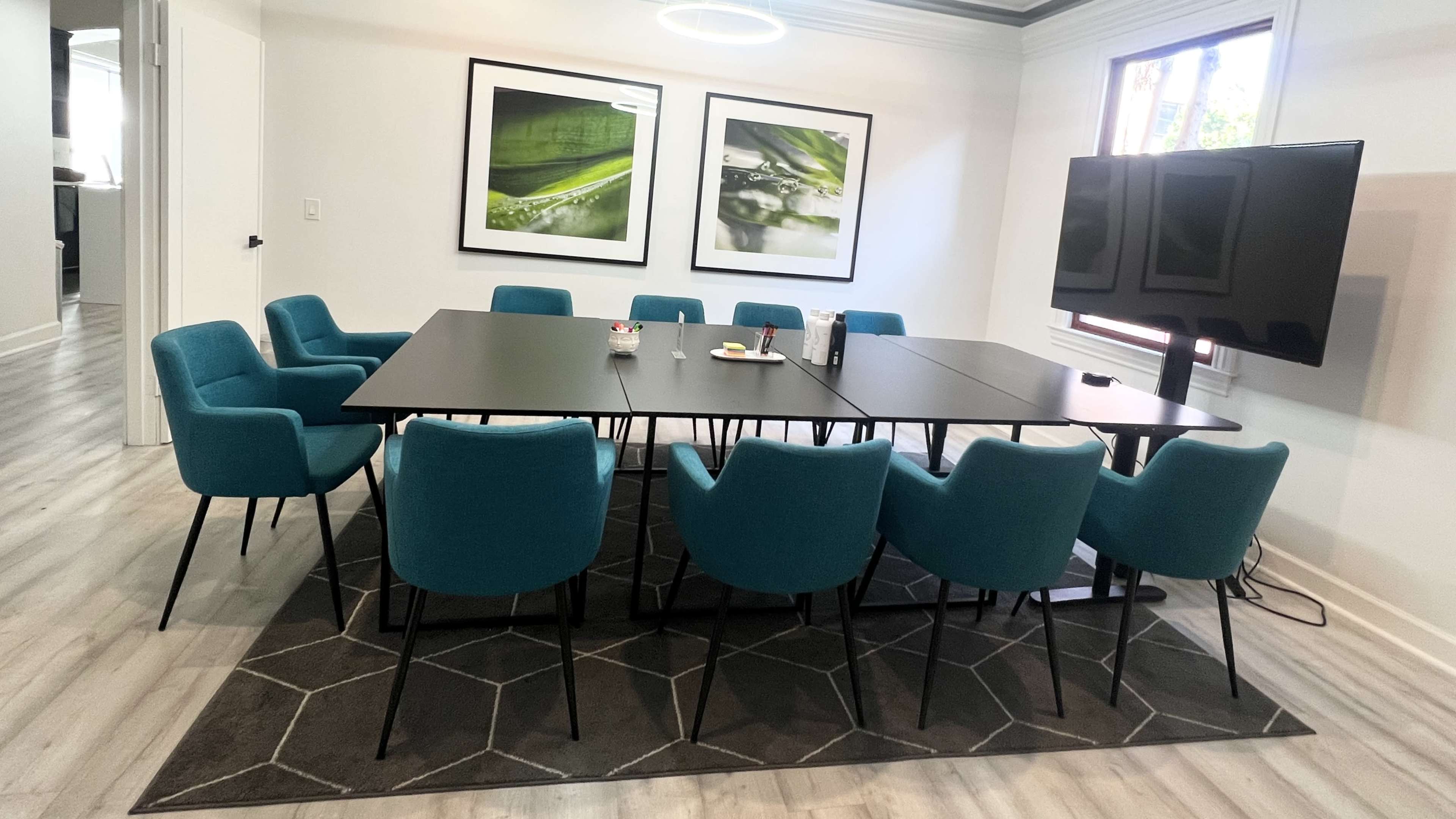 A modern conference room with a large rectangular table surrounded by twelve teal chairs, two framed green-themed pictures on the wall, and a wall-mounted television.