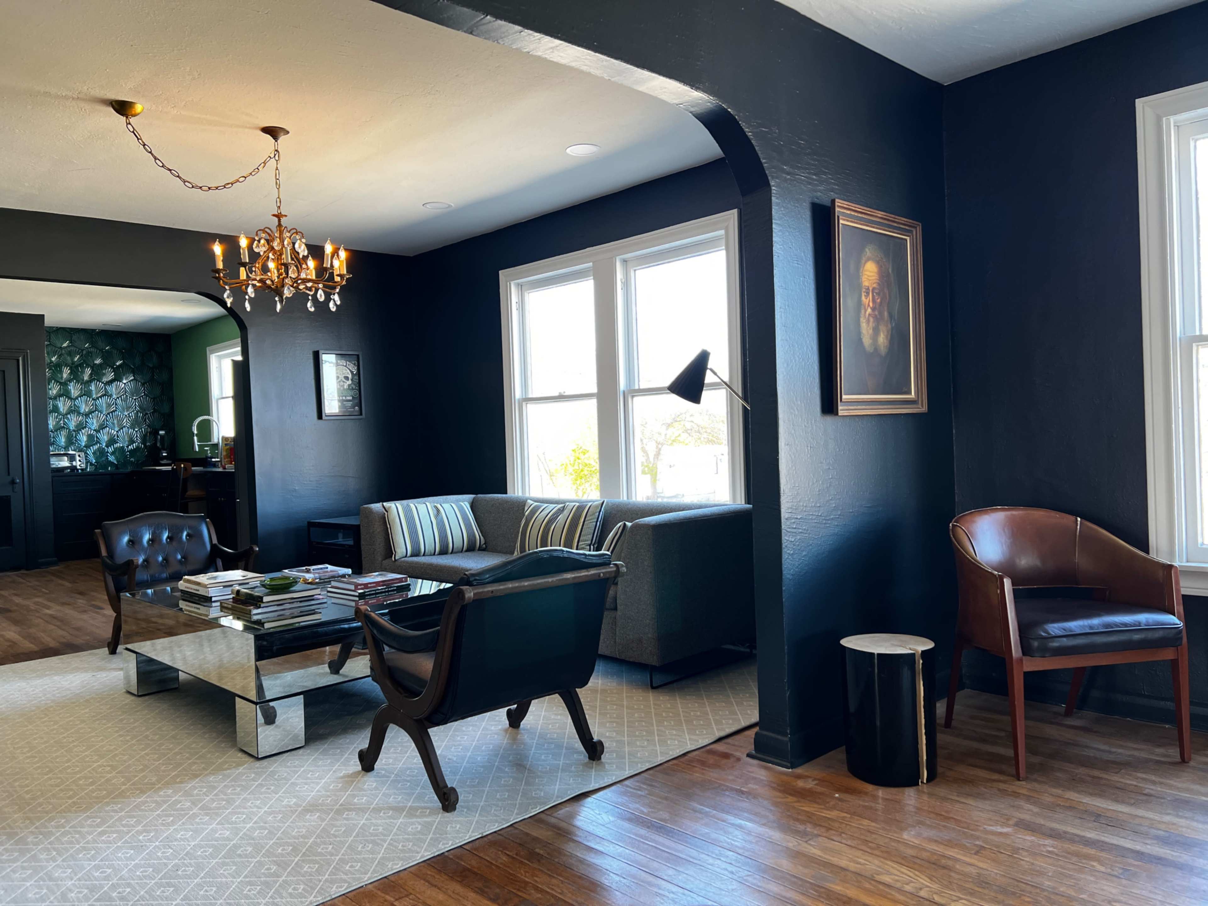 A modern living room with dark walls, a chandelier, a sectional sofa, a coffee table stacked with magazines, and a mix of armchairs.