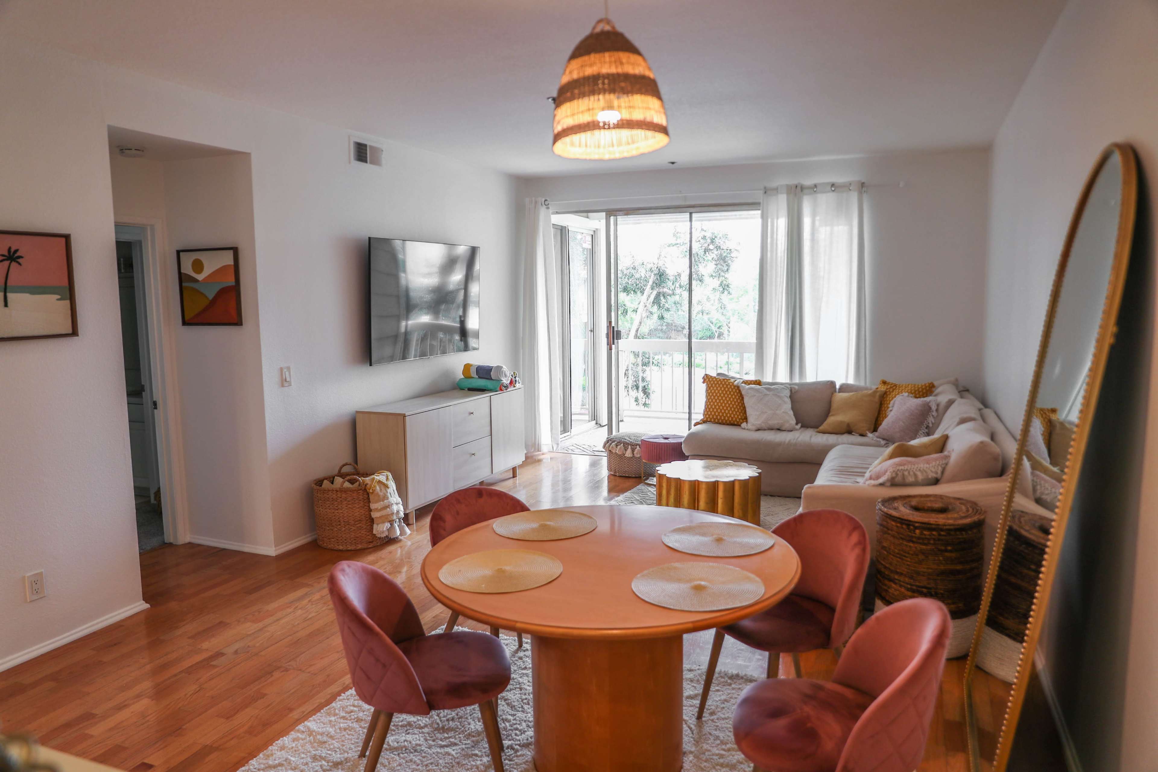 A modern living room features a light-colored sofa, a circular dining table with pink chairs, and large windows overlooking greenery.