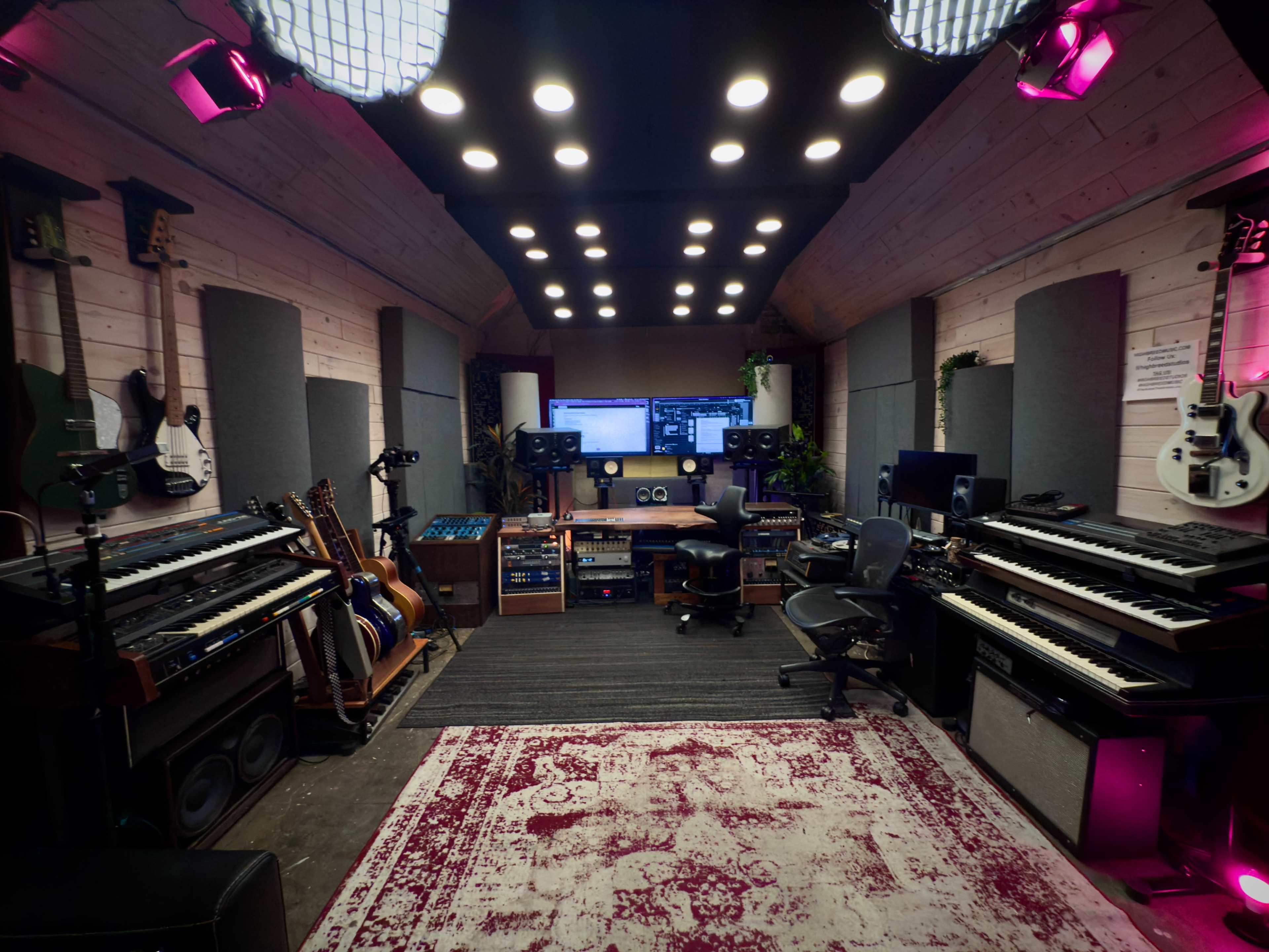 The image shows a spacious music studio equipped with various instruments, audio equipment, and a computer setup, illuminated by overhead lights.