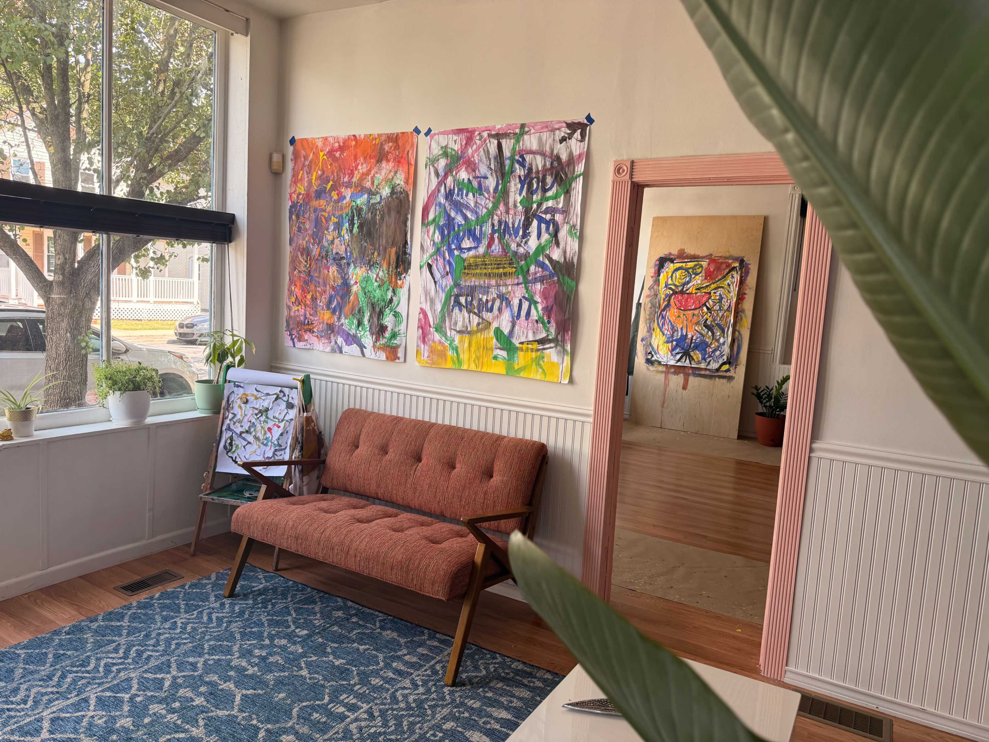 The image shows a bright room with a cozy orange couch, colorful abstract paintings on the walls, and a view into another space with additional artwork.