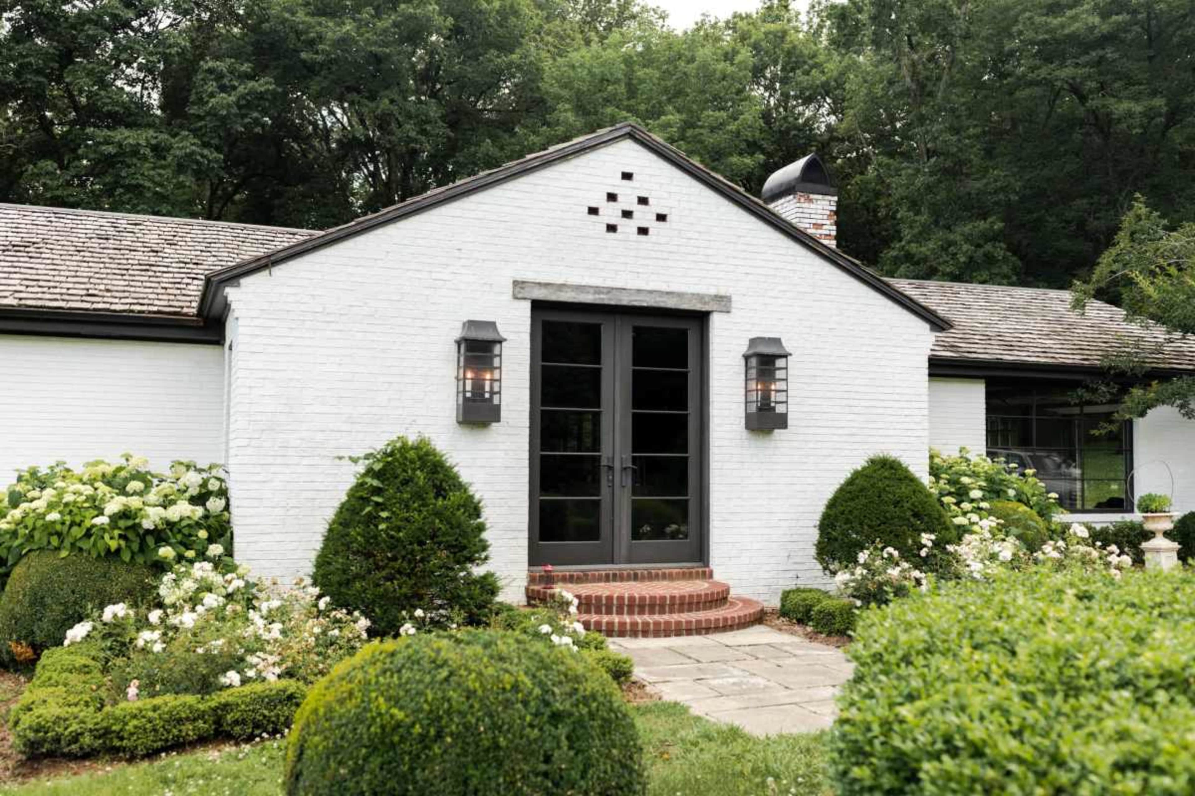 A white brick house features a sloped roof, symmetrical front garden with manicured shrubs, and a brick walkway leading to double black doors.
