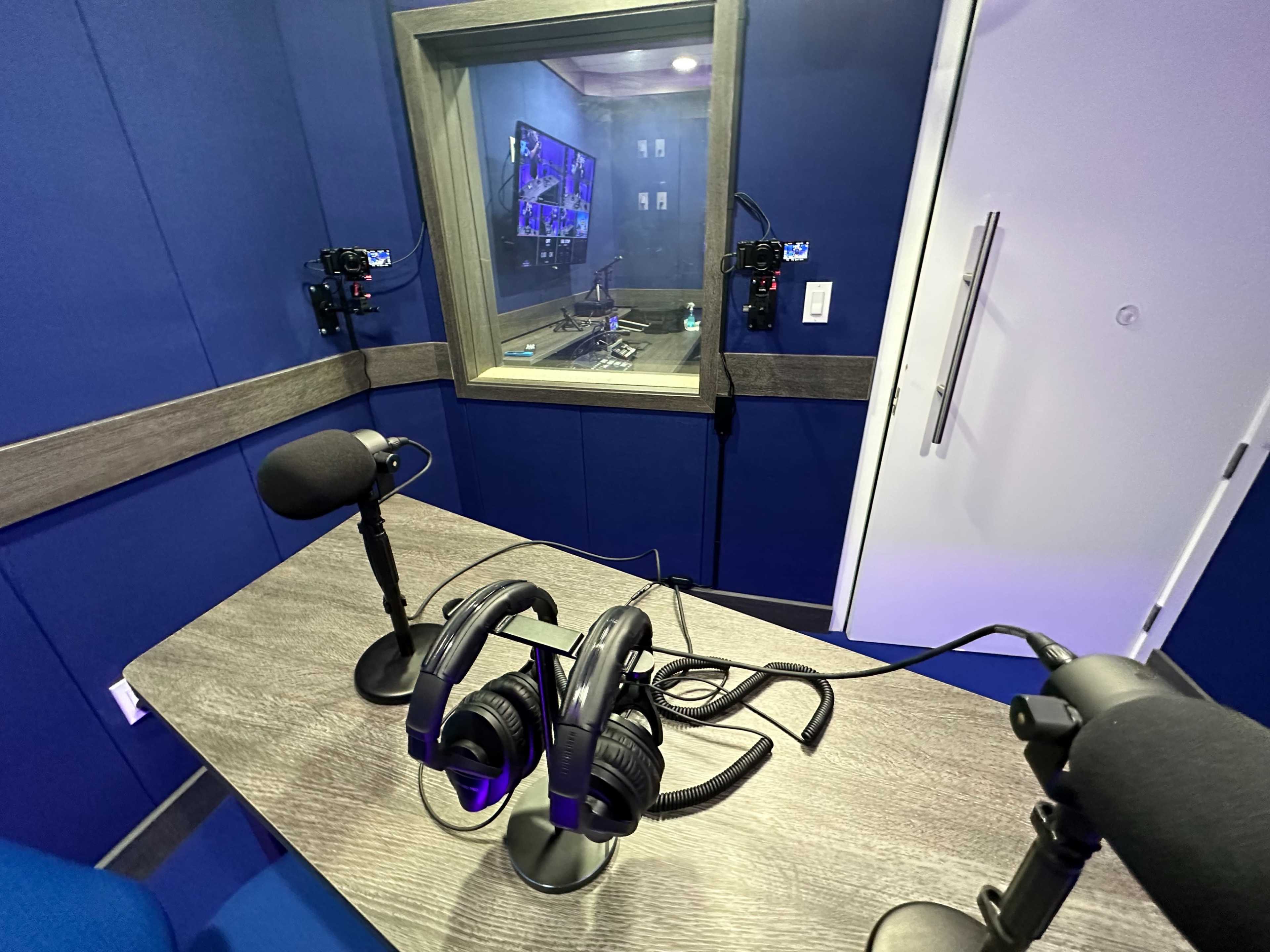 A recording studio with two microphones and headphones on a table, and a soundproof booth visible in the background.