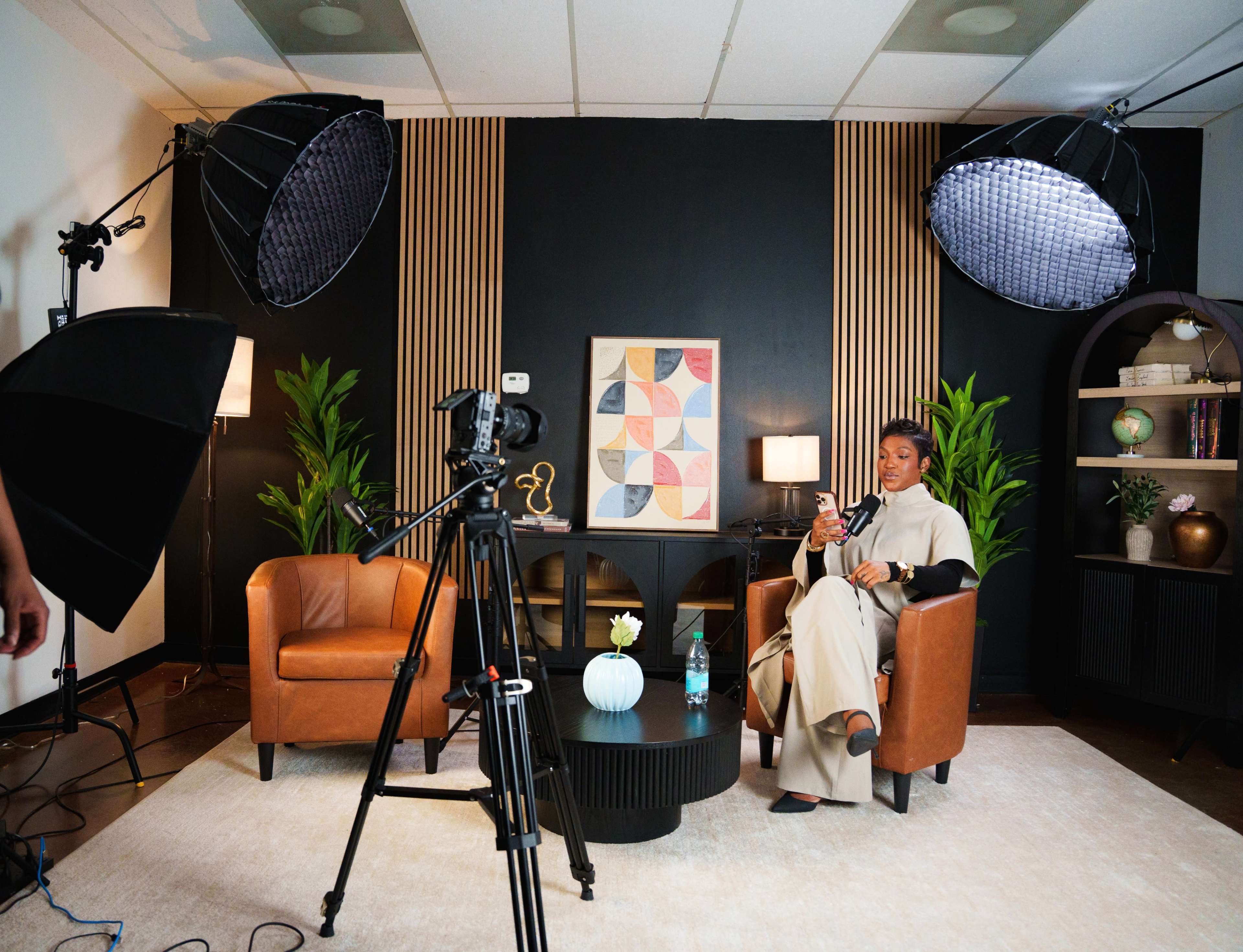 A well-lit studio setup features two brown armchairs, a round black coffee table, and a woman seated in one chair, holding a phone, with studio lights and a camera in view.