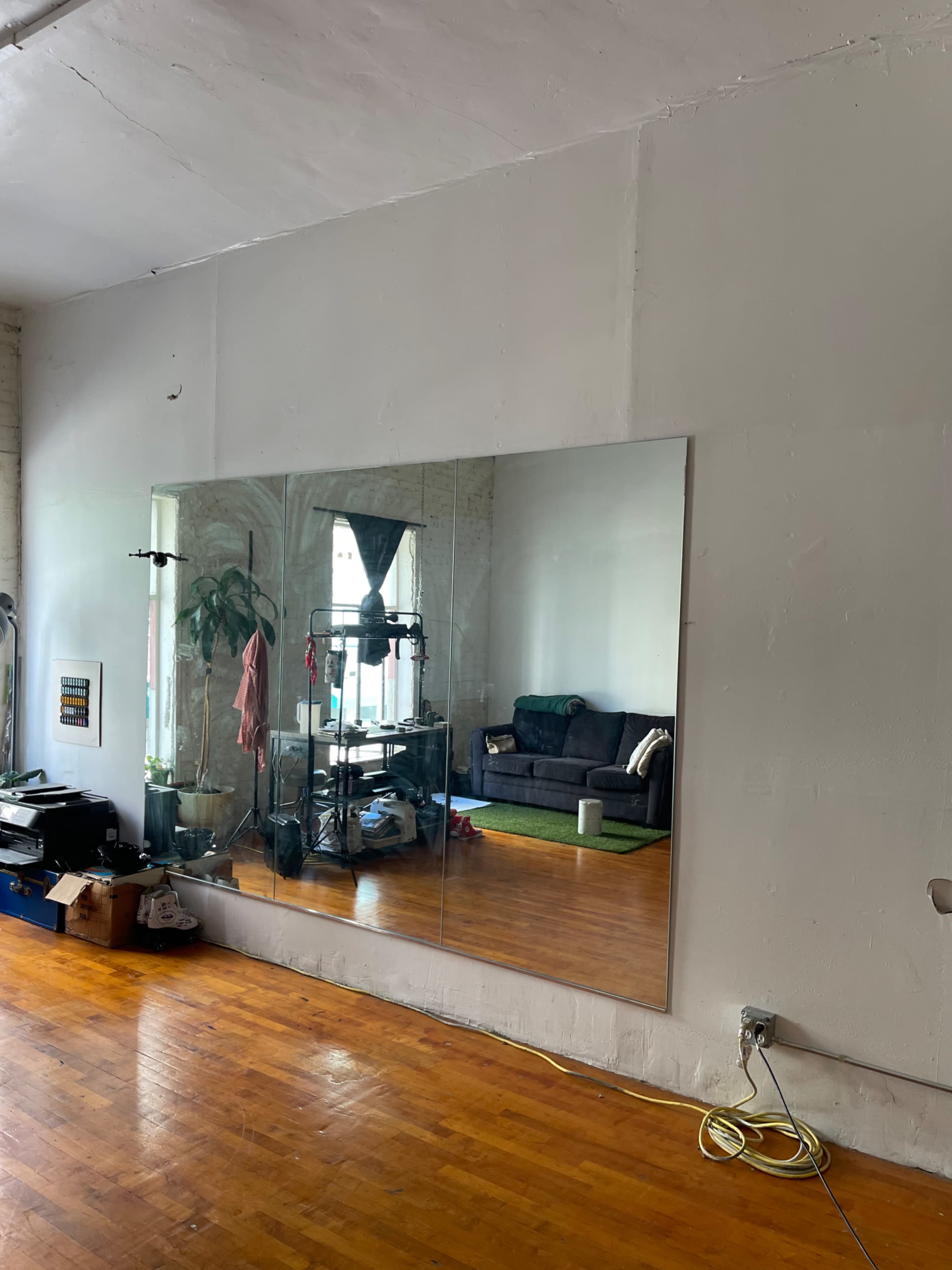 A large mirror is mounted on a wall in a room with a wooden floor, showing a glimpse of a couch and plants in the reflection.
