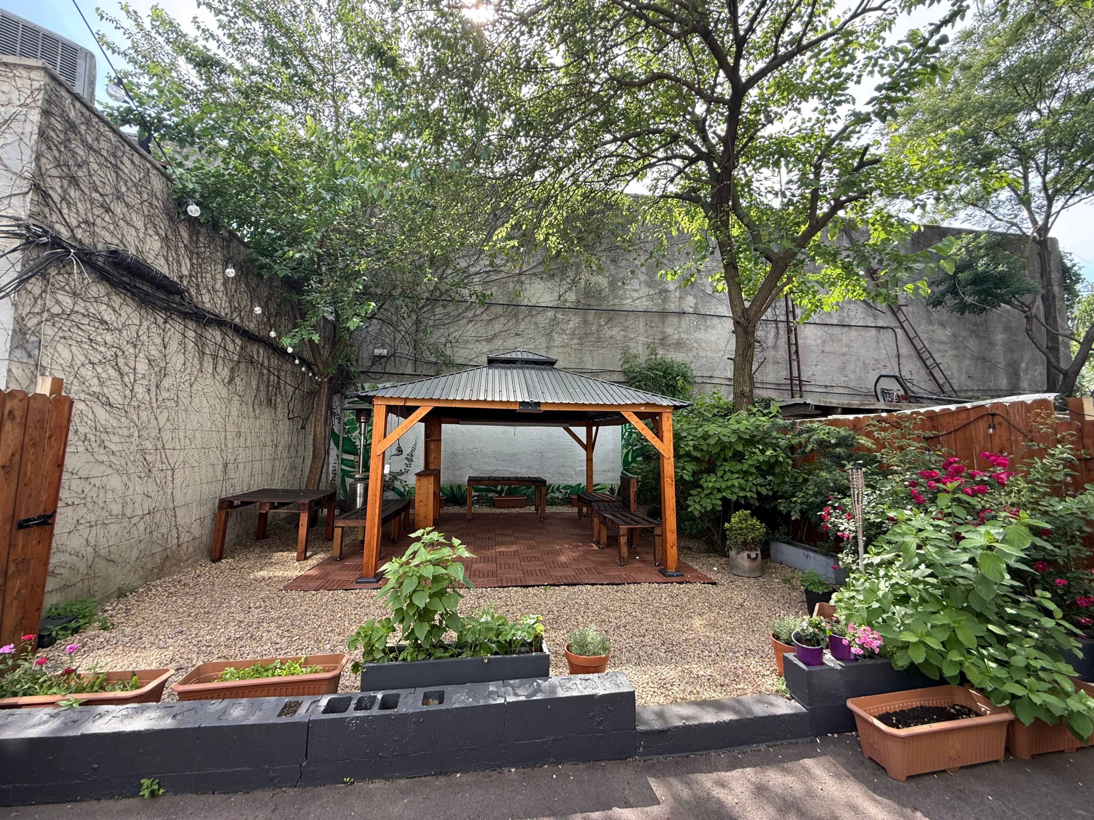 The image shows a shaded outdoor seating area with a wooden gazebo surrounded by plants and gravel.