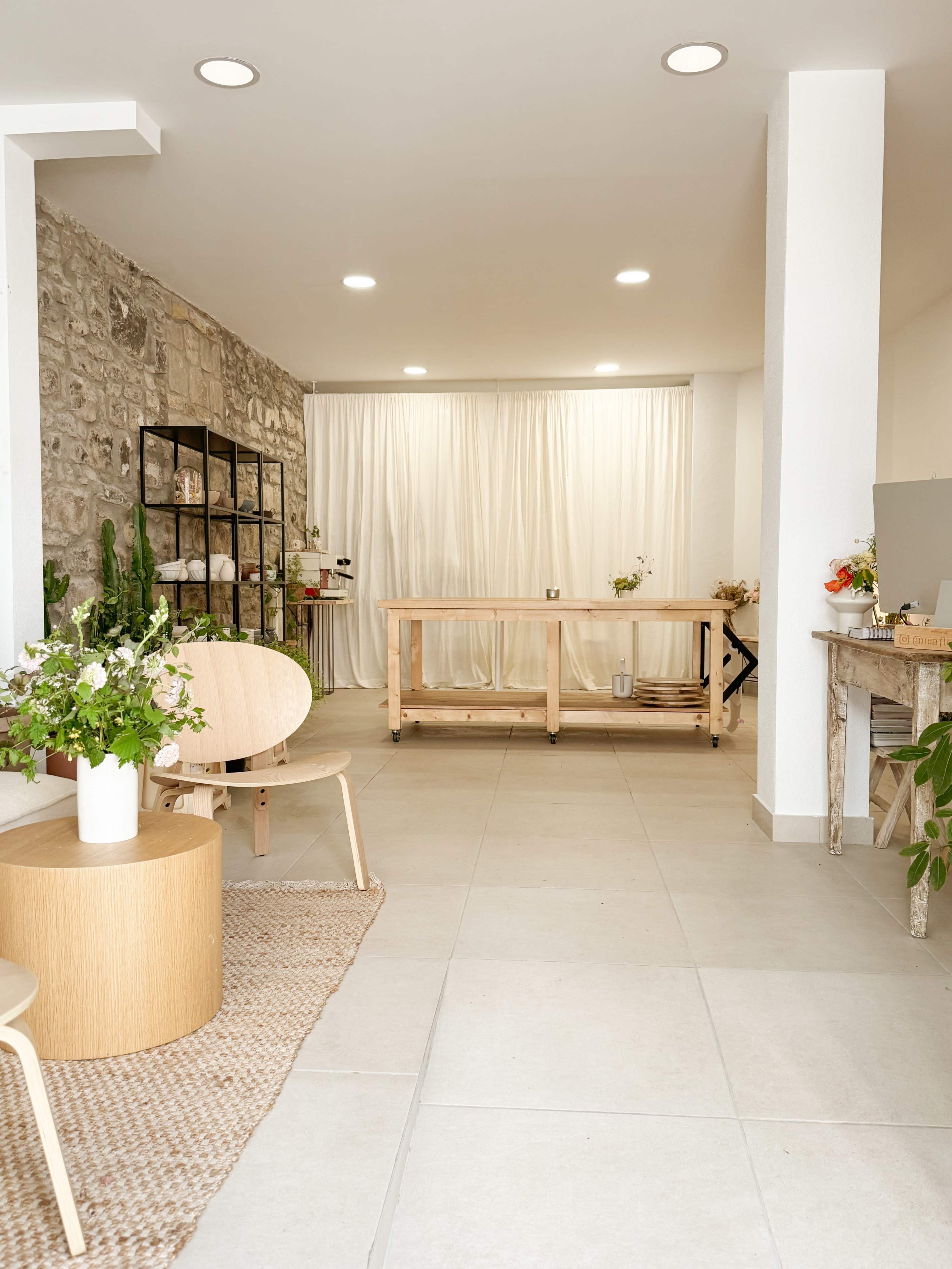 The image shows a minimalist interior space with a wooden table, light-colored flooring, and a stone wall, featuring plants and soft curtains in the background.