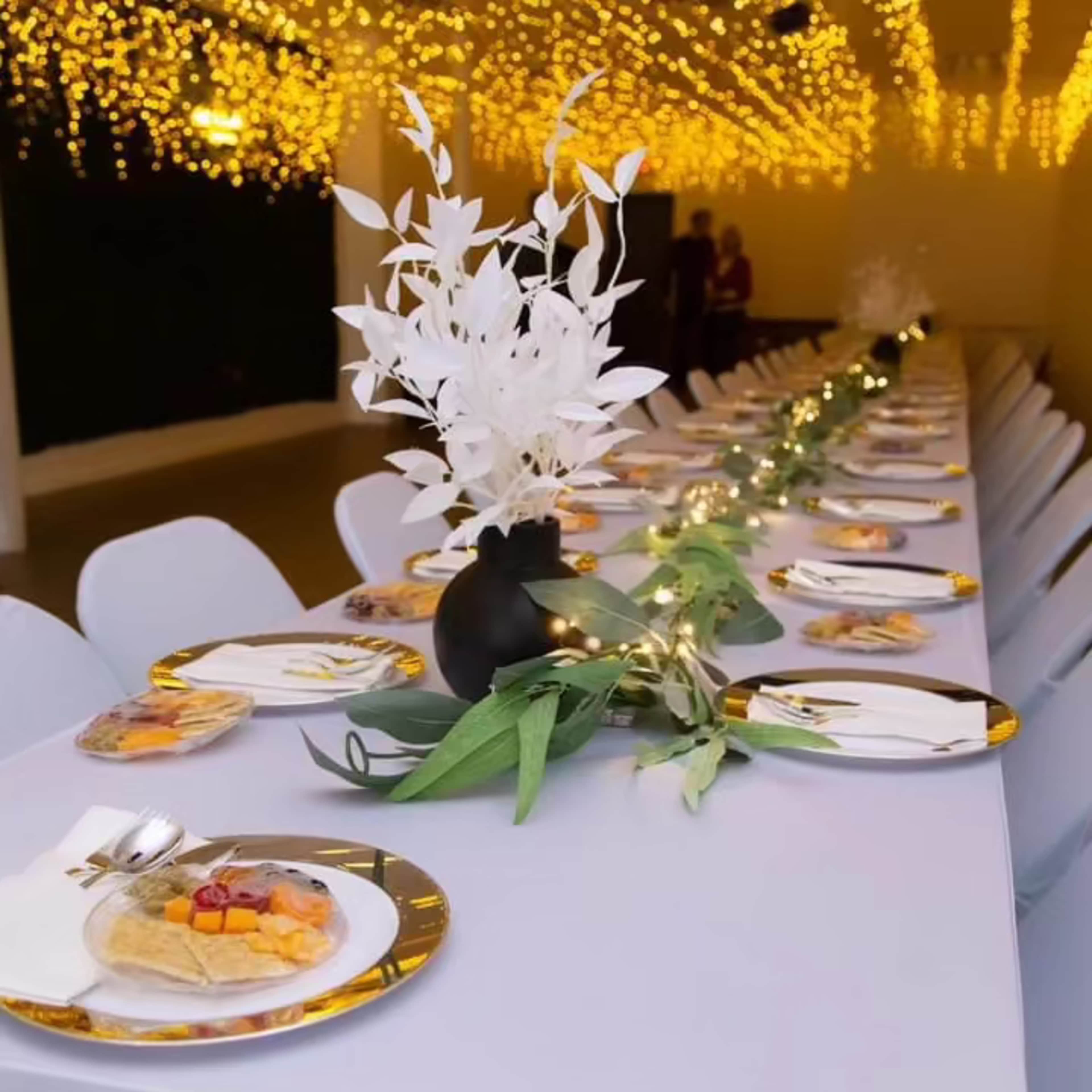 A long dining table is elegantly set with white plates and a decorative vase of white foliage, surrounded by soft lighting from overhead strings of lights.