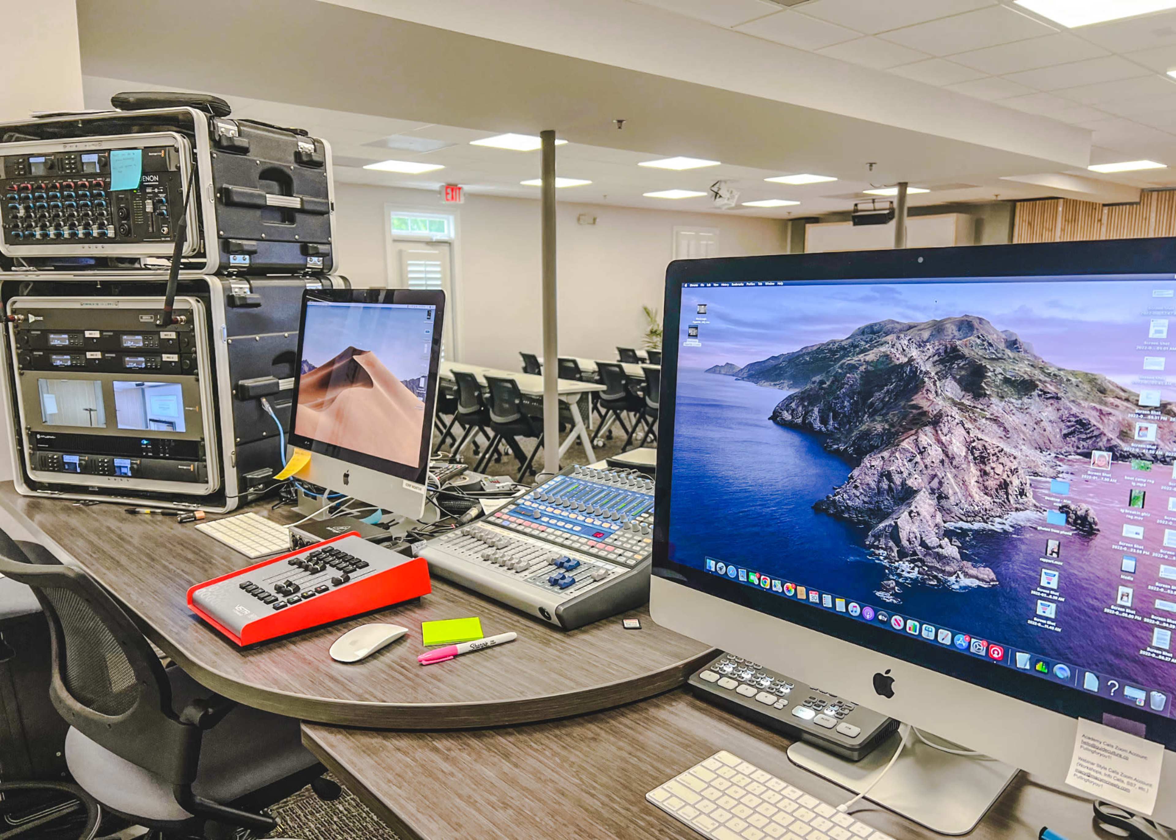 A workspace with multiple computers, audio equipment, and a mixing console set up in a room with chairs arranged for an event.