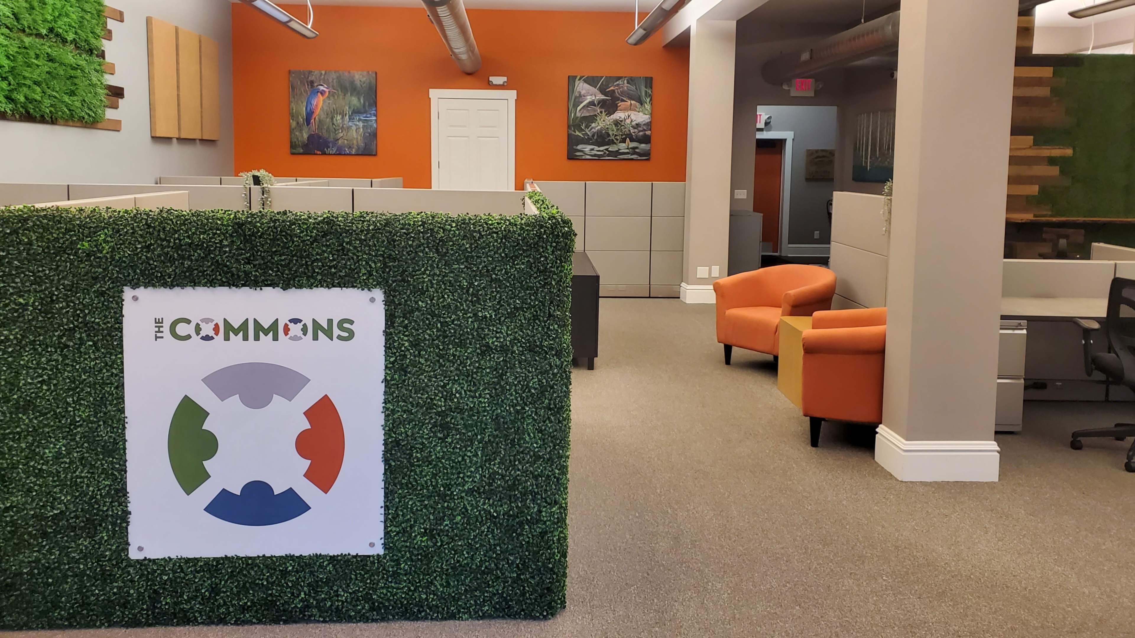 An office space with gray cubicles, orange walls featuring artwork, and a cozy seating area with two orange chairs.