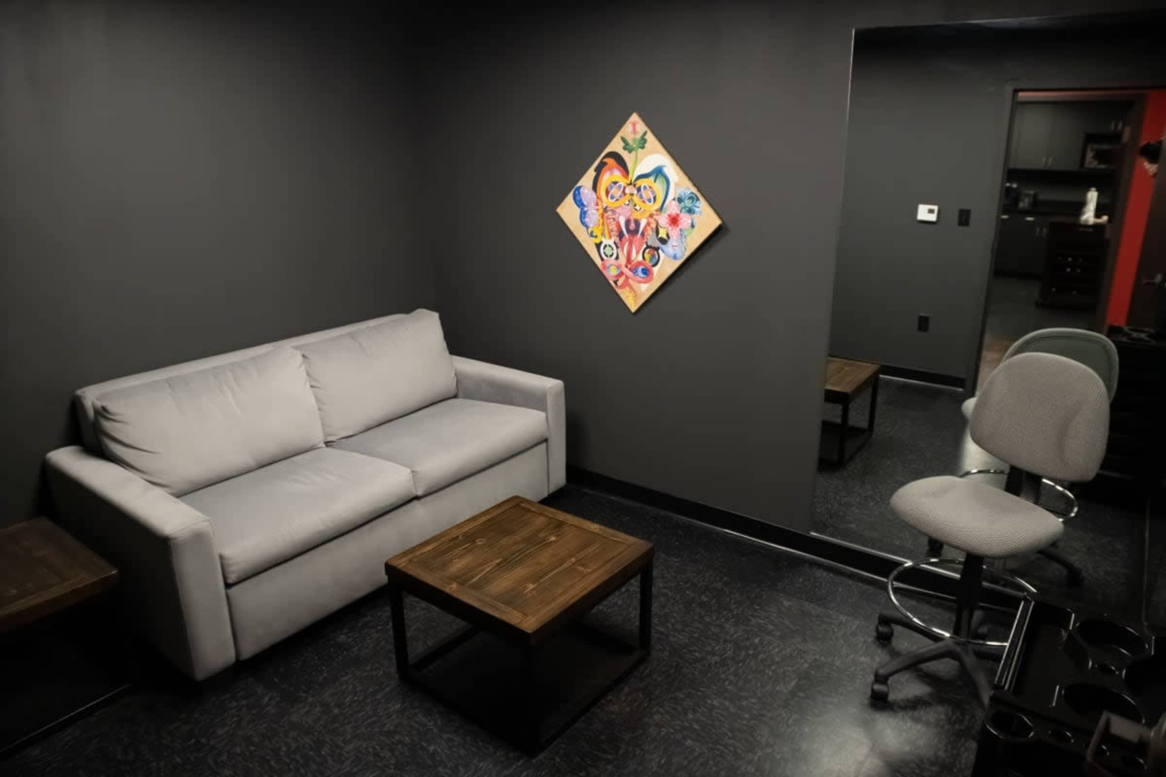 A small, dark room with a gray sofa, a wooden table, a swivel chair, and a colorful painting on the wall.
