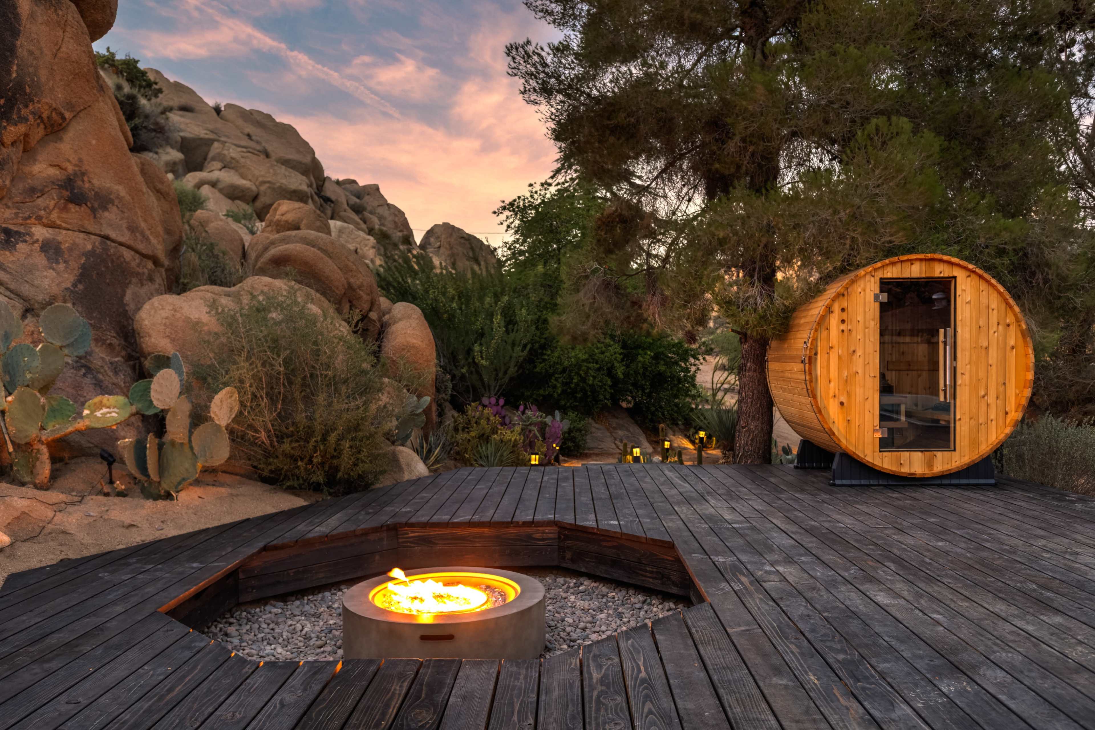 A wooden deck features a circular fire pit surrounded by rocks, with a sauna-shaped structure and desert vegetation in the background.