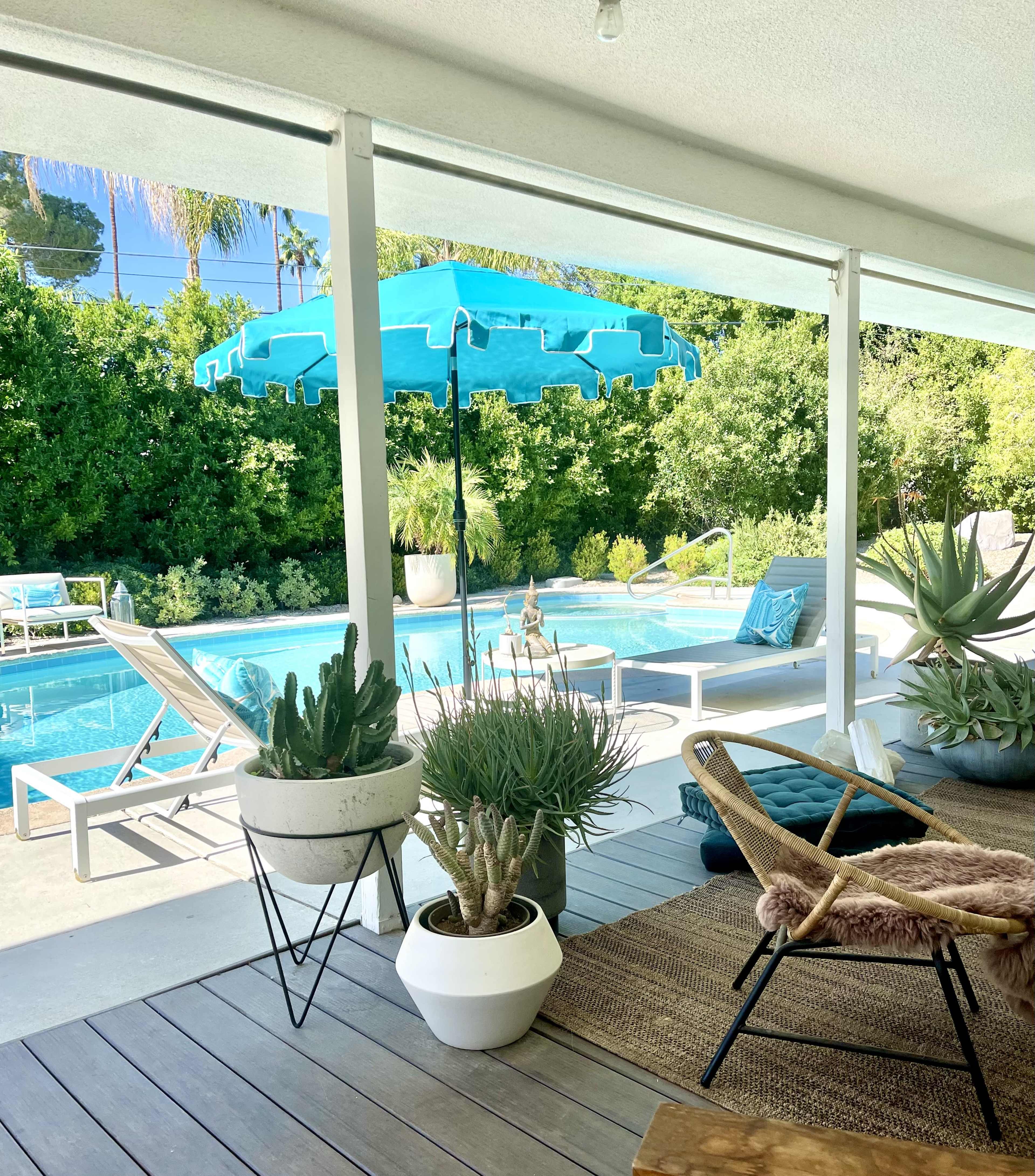 A covered patio overlooks a swimming pool surrounded by greenery, with potted plants and lounge chairs arranged nearby.