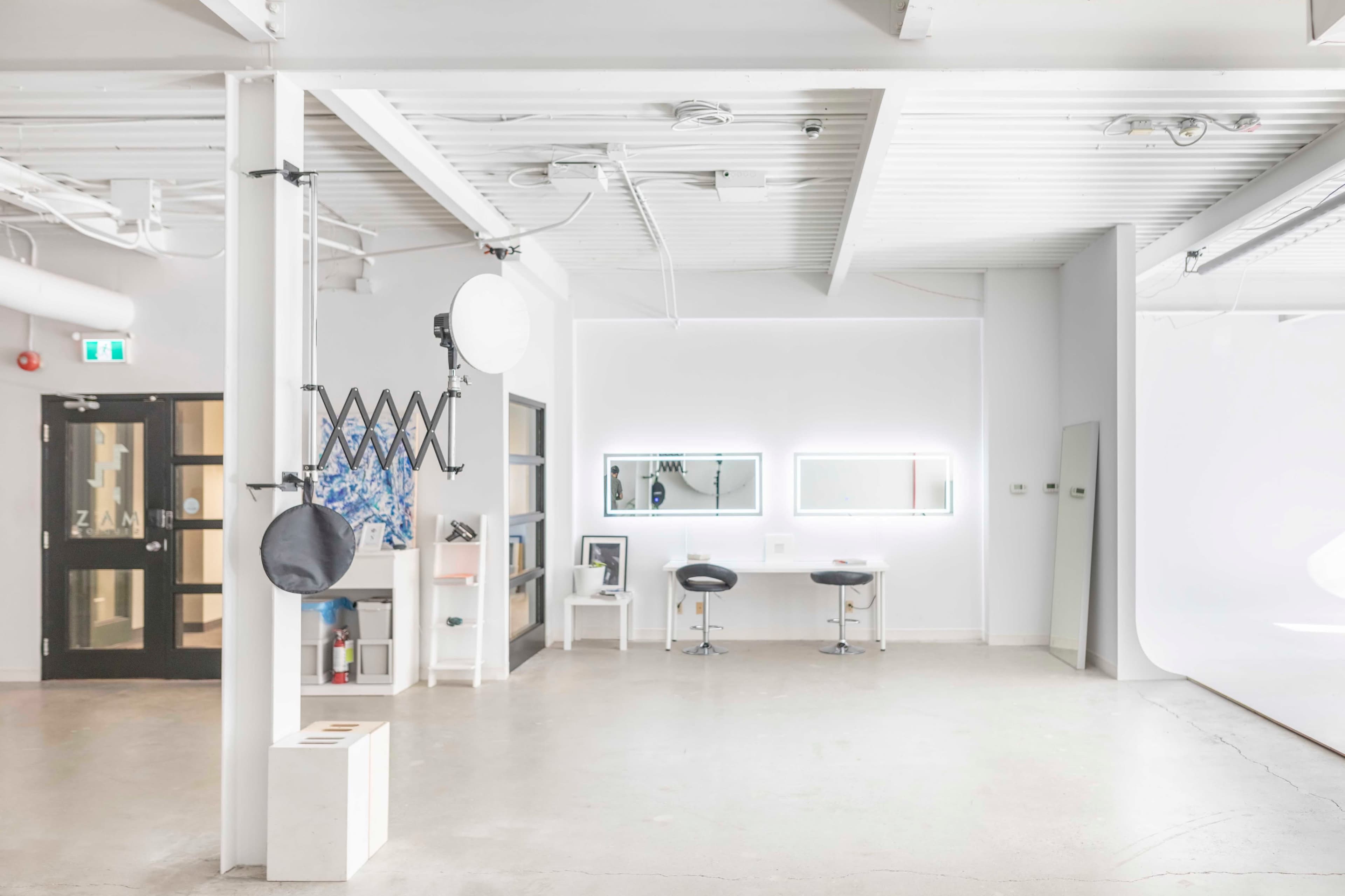A spacious studio with white walls, a concrete floor, and a few pieces of furniture, including two black chairs and a mirror setup.