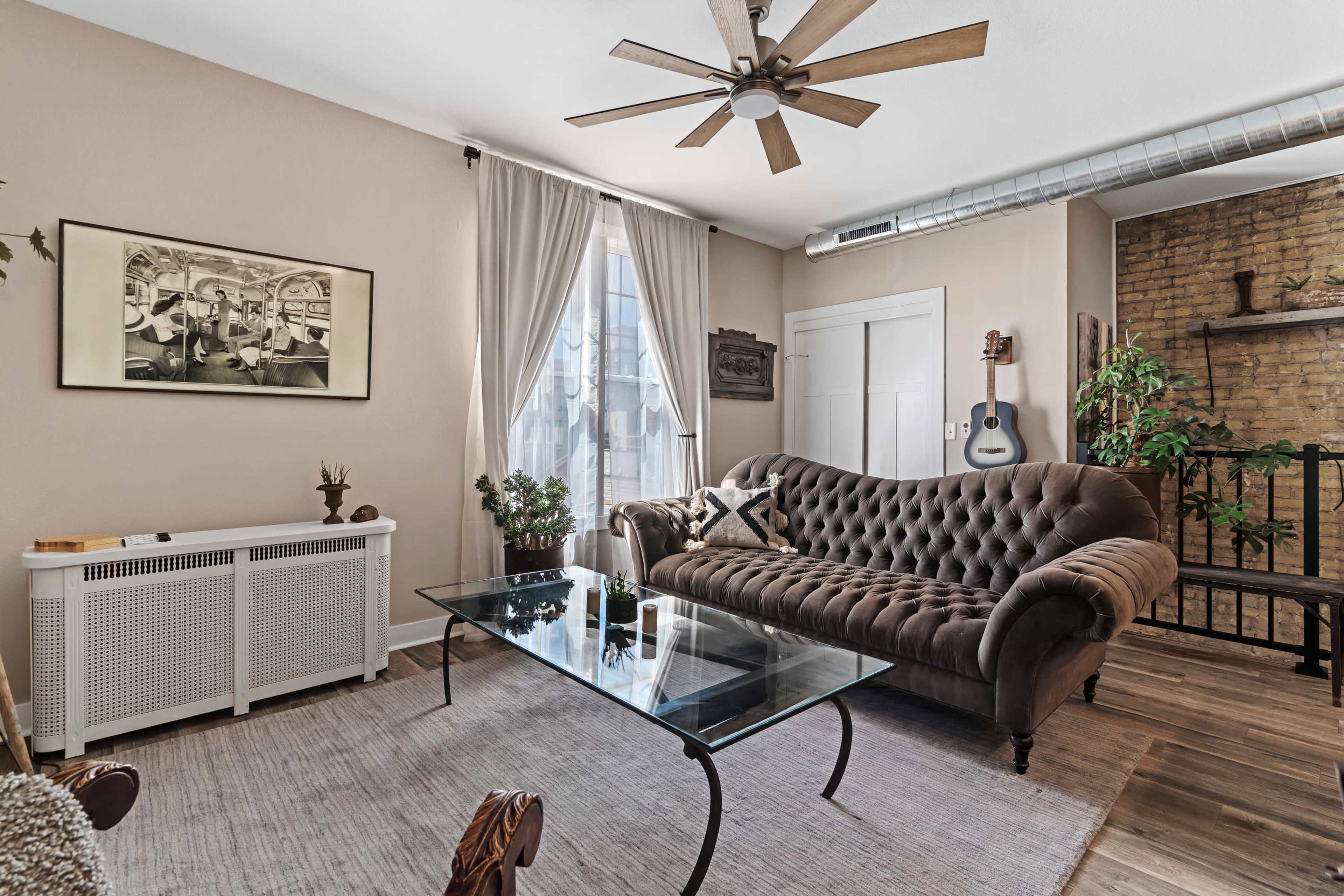 A living room featuring a brown tufted sofa, a glass coffee table, and a mix of natural plants, with a fan overhead and a guitar leaning against the wall.