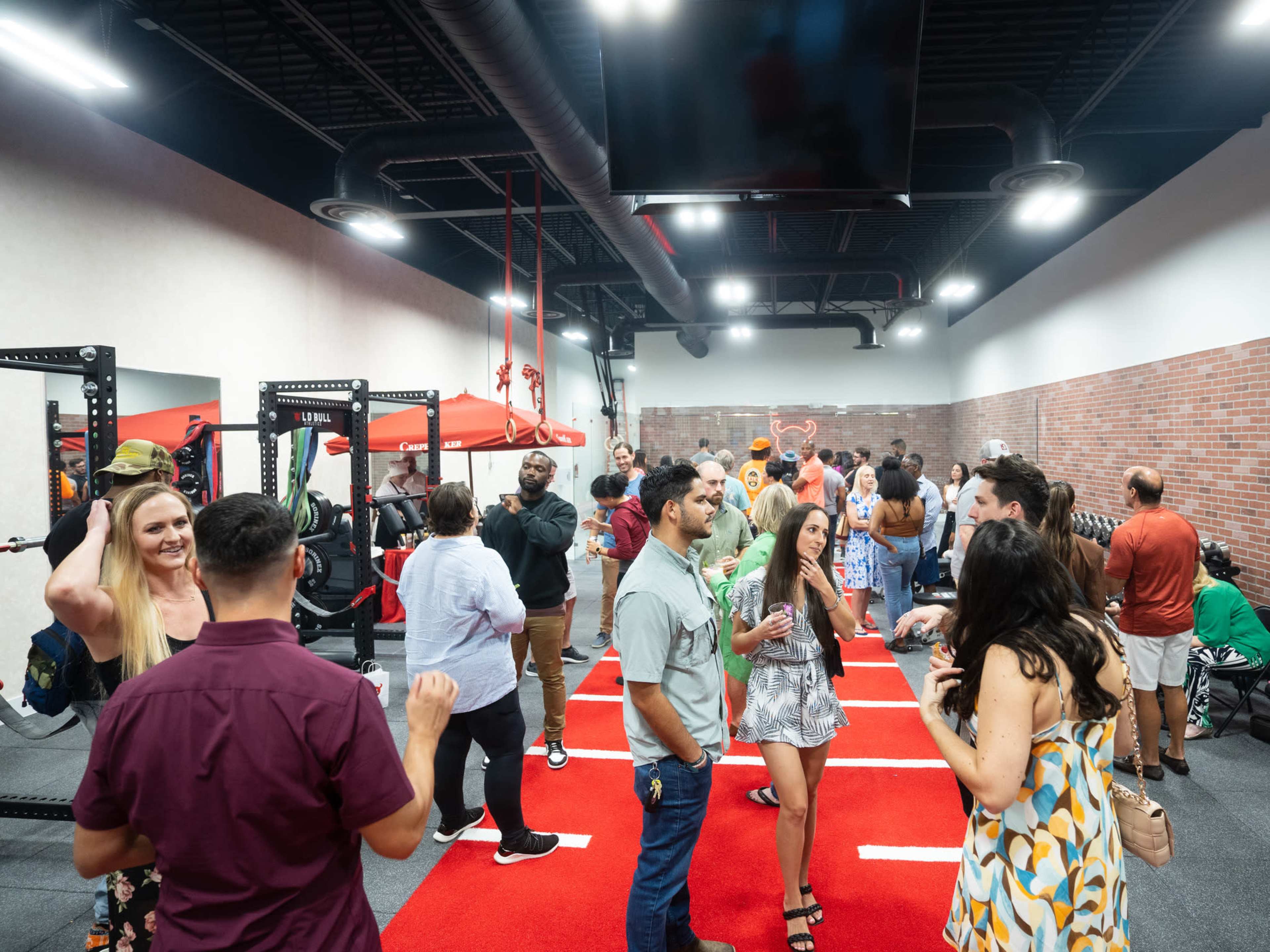 A group of people socializes in a gym setting with fitness equipment and red flooring.