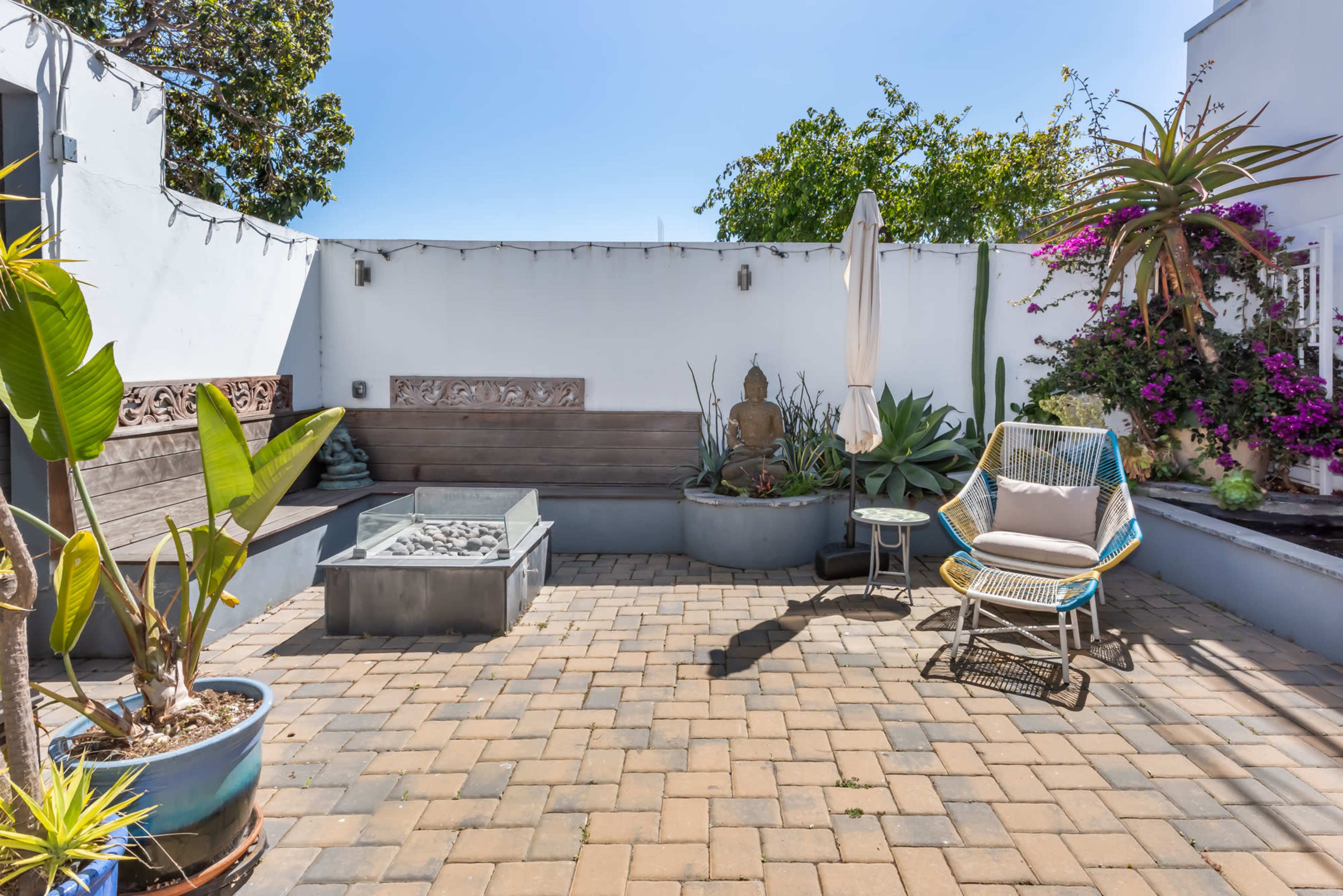 A landscaped outdoor patio featuring paver stones, a seating area, a fire pit, and decorative plants.