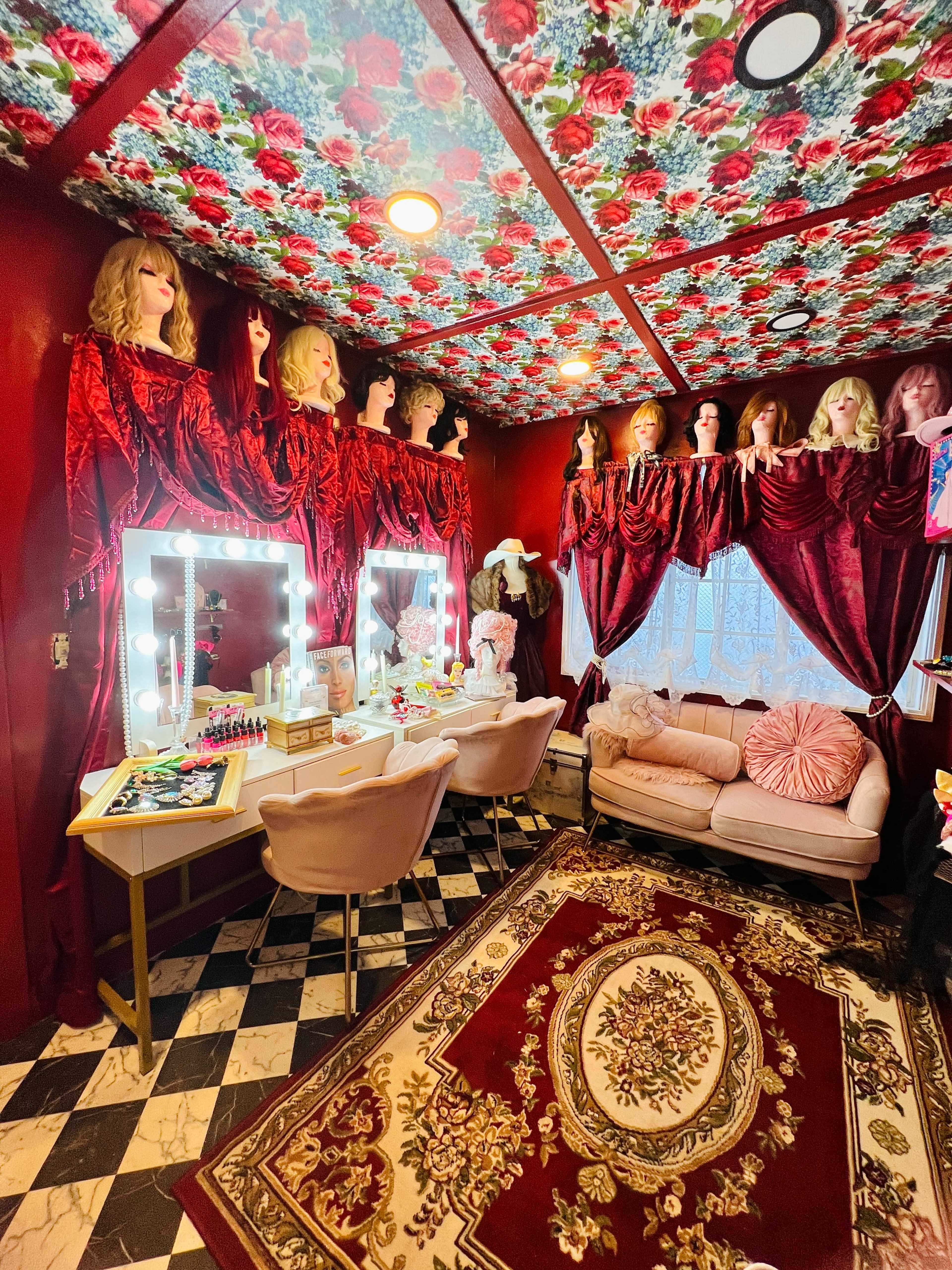 A small room decorated with floral wallpaper, adorned with mannequin heads, velvet curtains, a lighted vanity, and a plush sofa on a patterned rug.