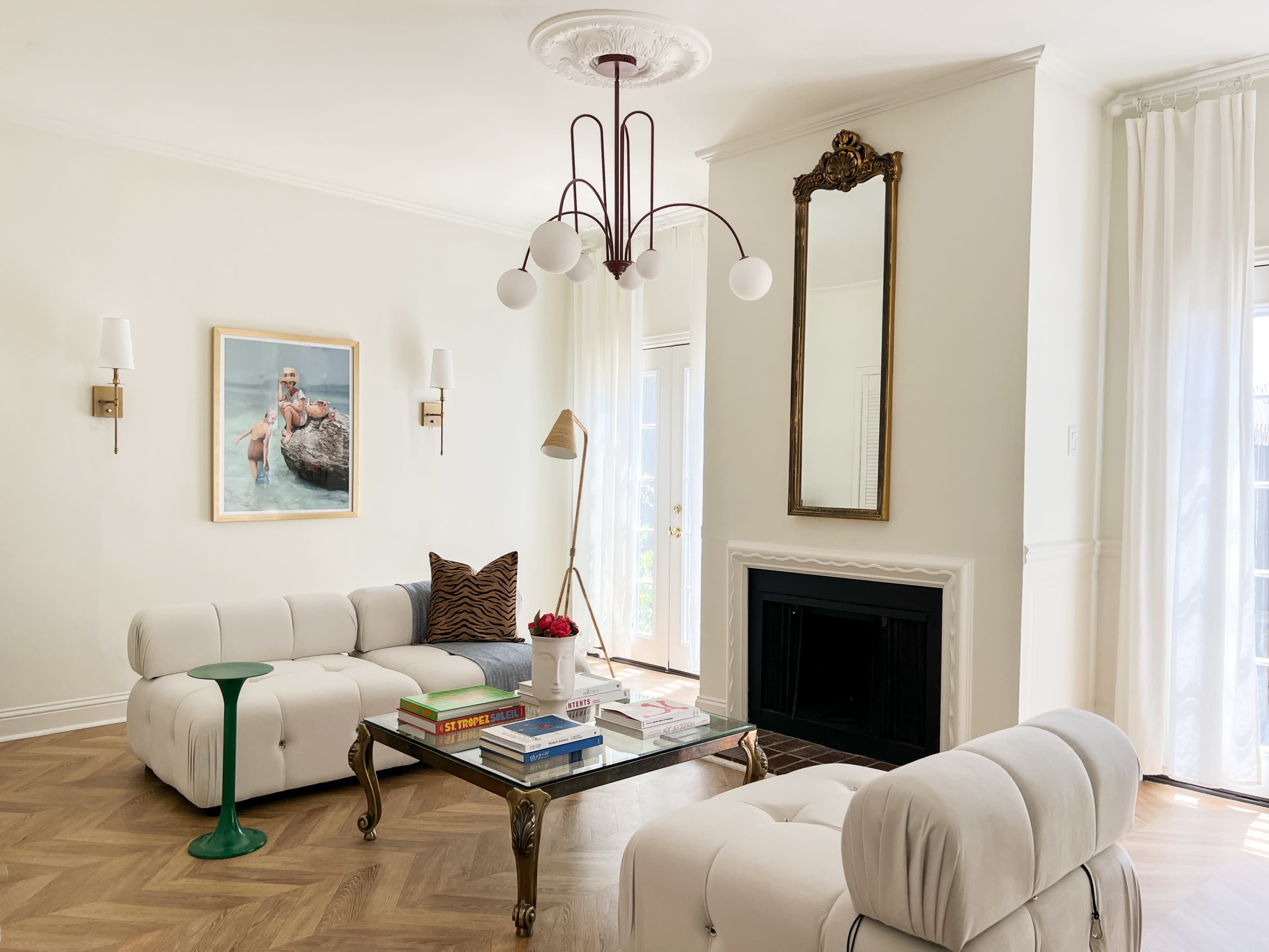 The living room features a light-colored sofa, a coffee table with books, a decorative mirror, and a large artwork on the wall, with floor-to-ceiling curtains and a fireplace.