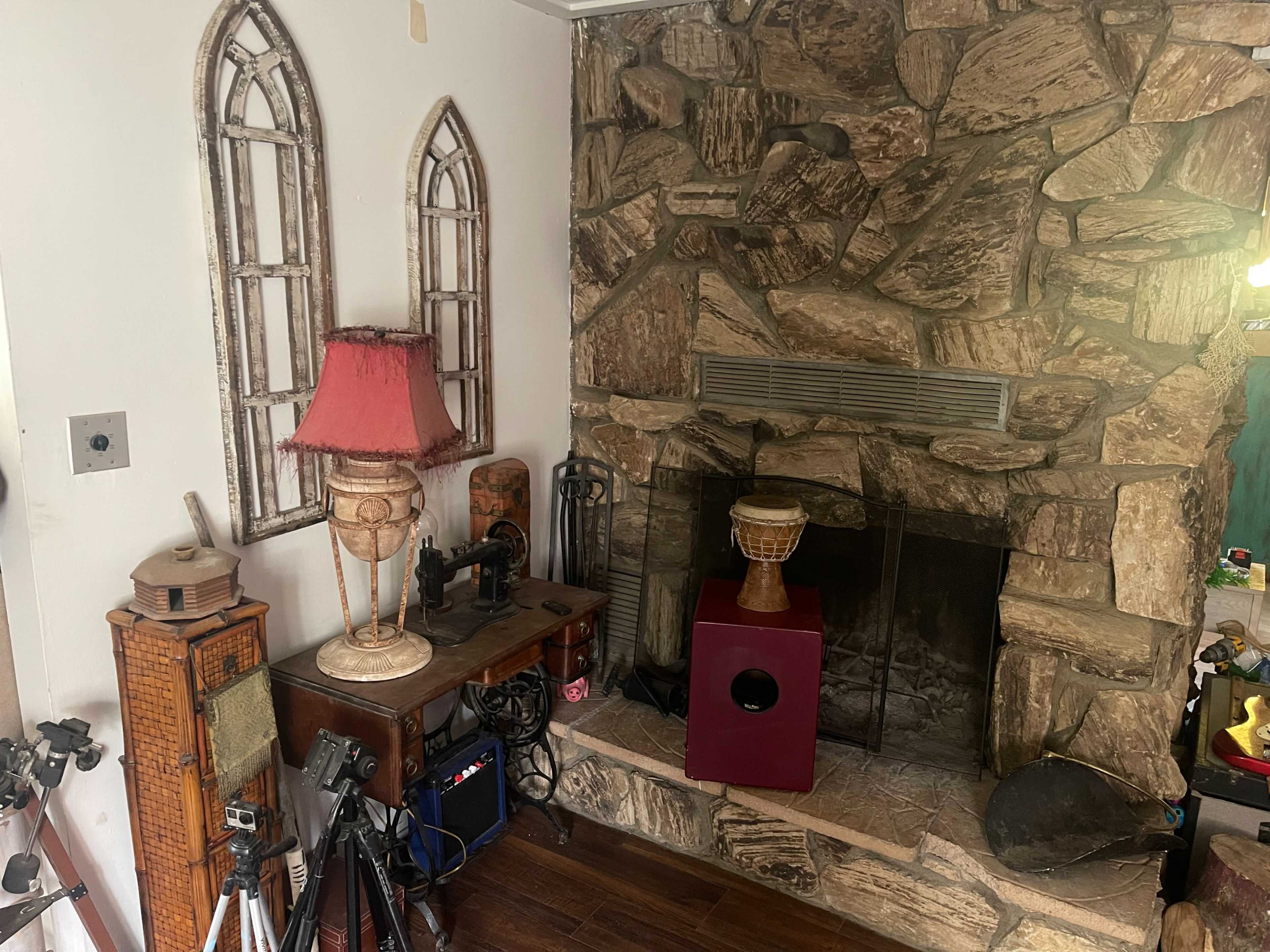 The image shows a living room corner featuring a stone fireplace, a wooden table with various objects, and a lamp alongside decorative window frames.