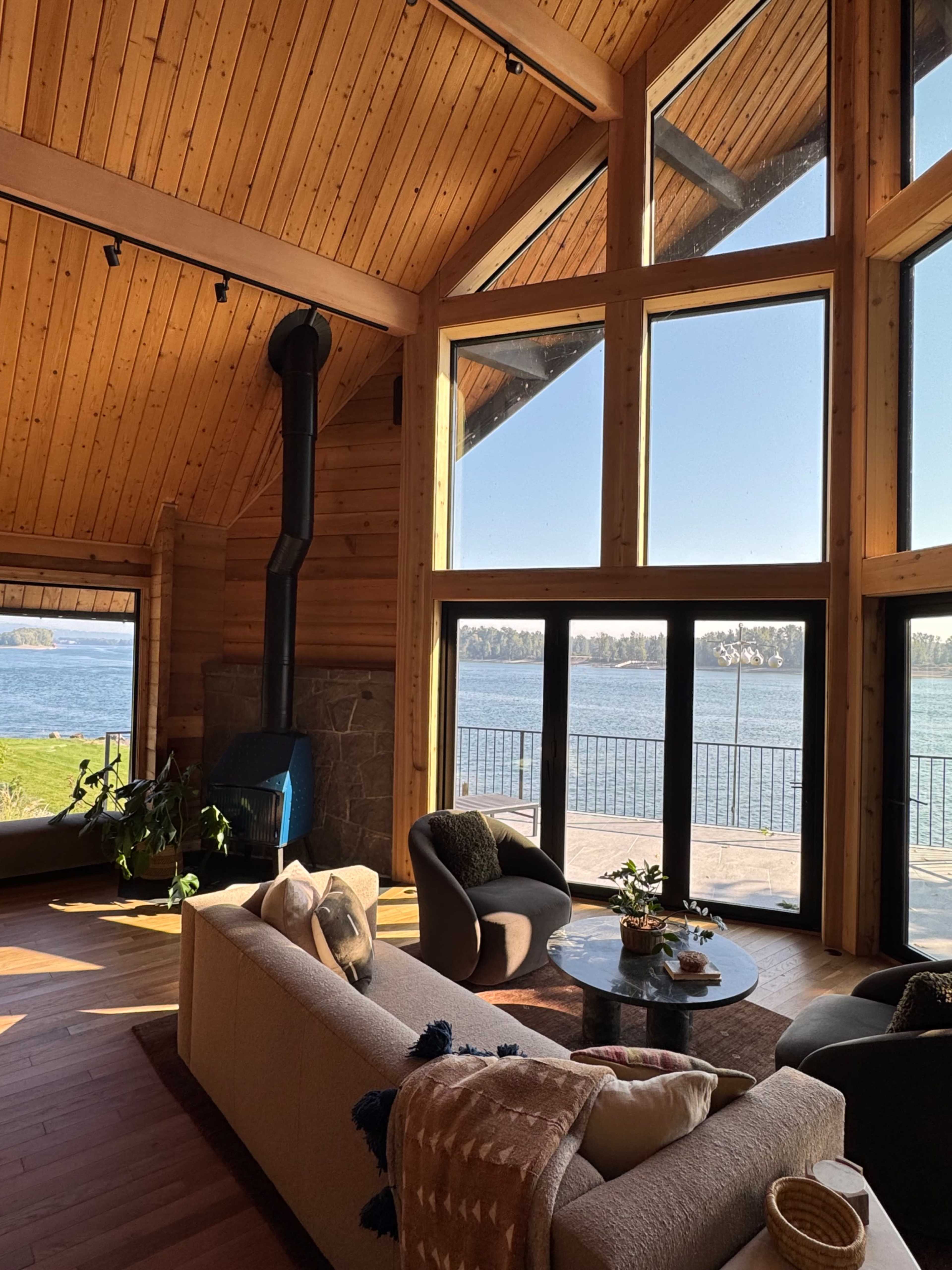 A modern living room with a large window showcasing a lakeside view, featuring wooden paneling, a sofa, and a wood stove.