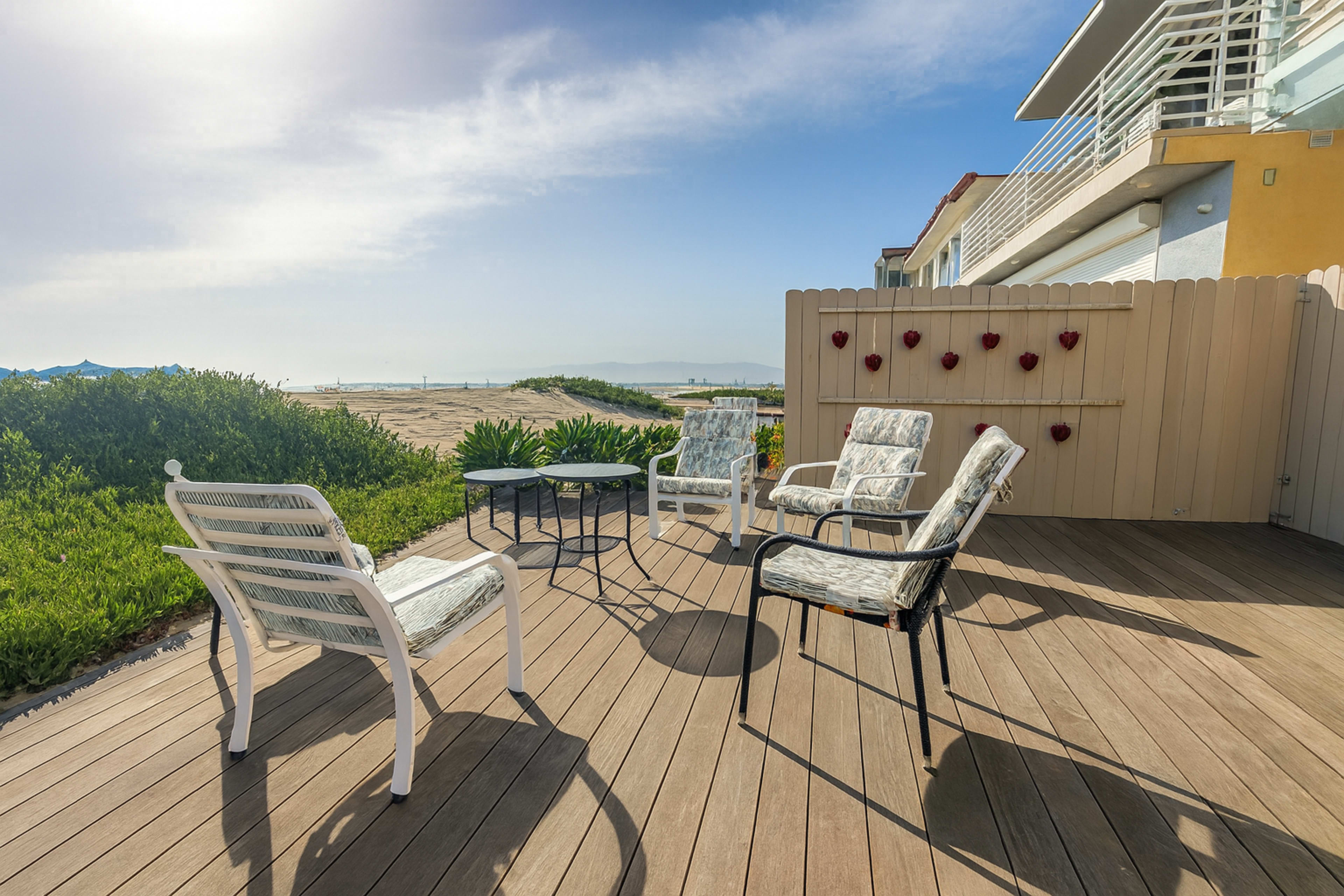 A patio area features four chairs arranged around a circular table, with a backdrop of greenery and a view of the water.