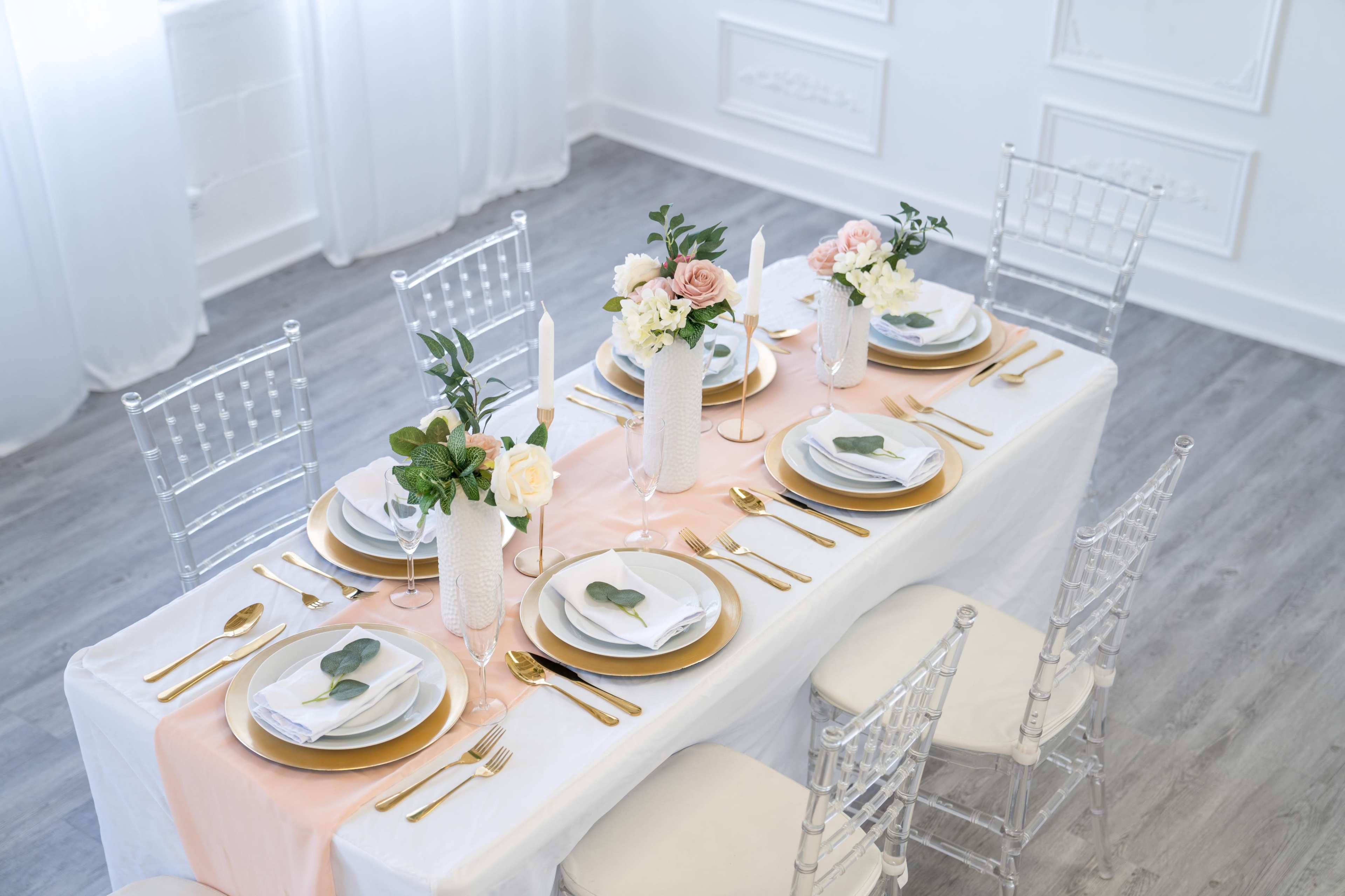 A beautifully set dining table with elegant floral arrangements, golden tableware, and a soft peach and white table runner in a bright, modern space.