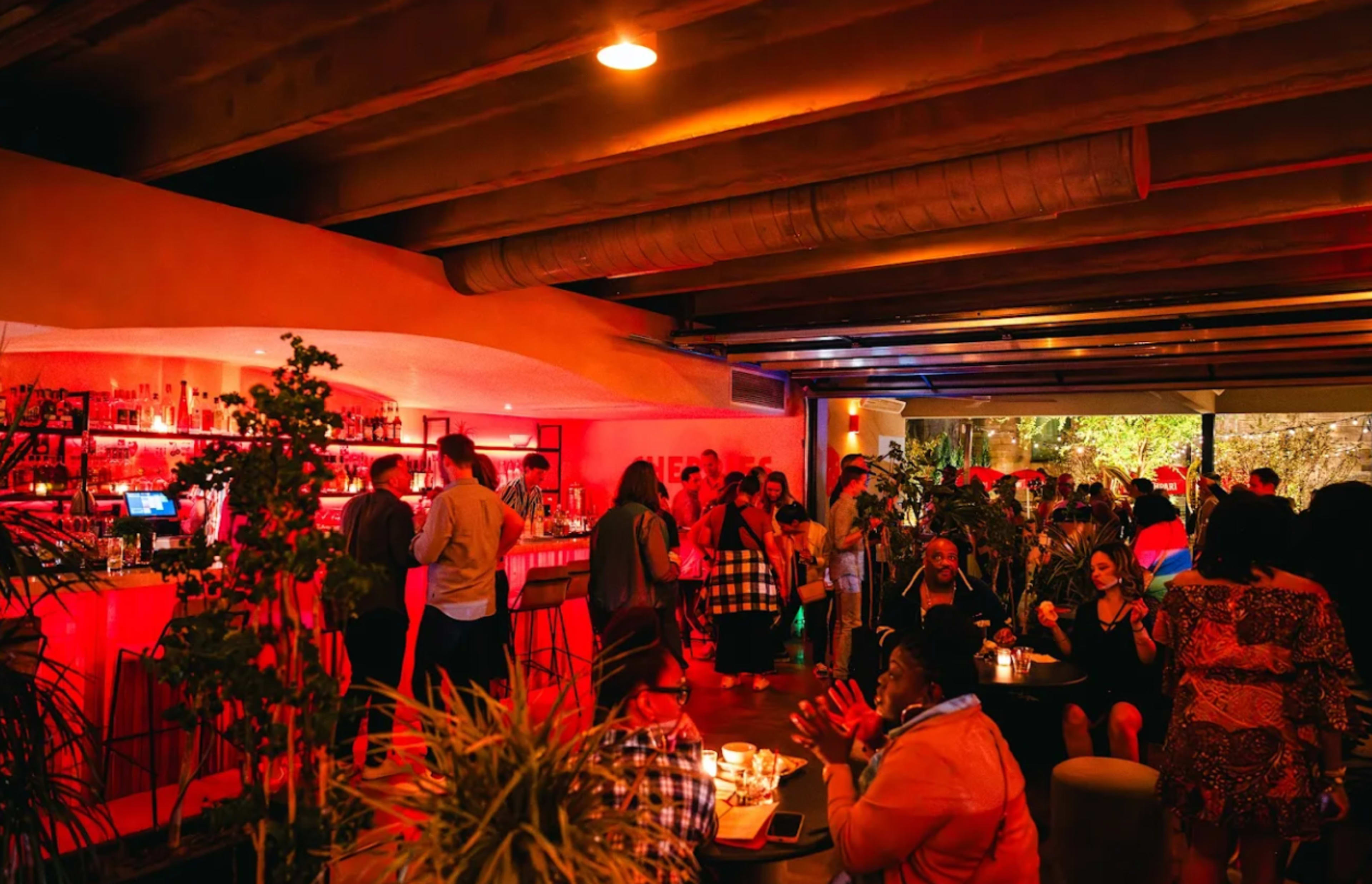 The image shows a lively bar interior filled with people socializing, illuminated by red lighting and surrounded by plants.