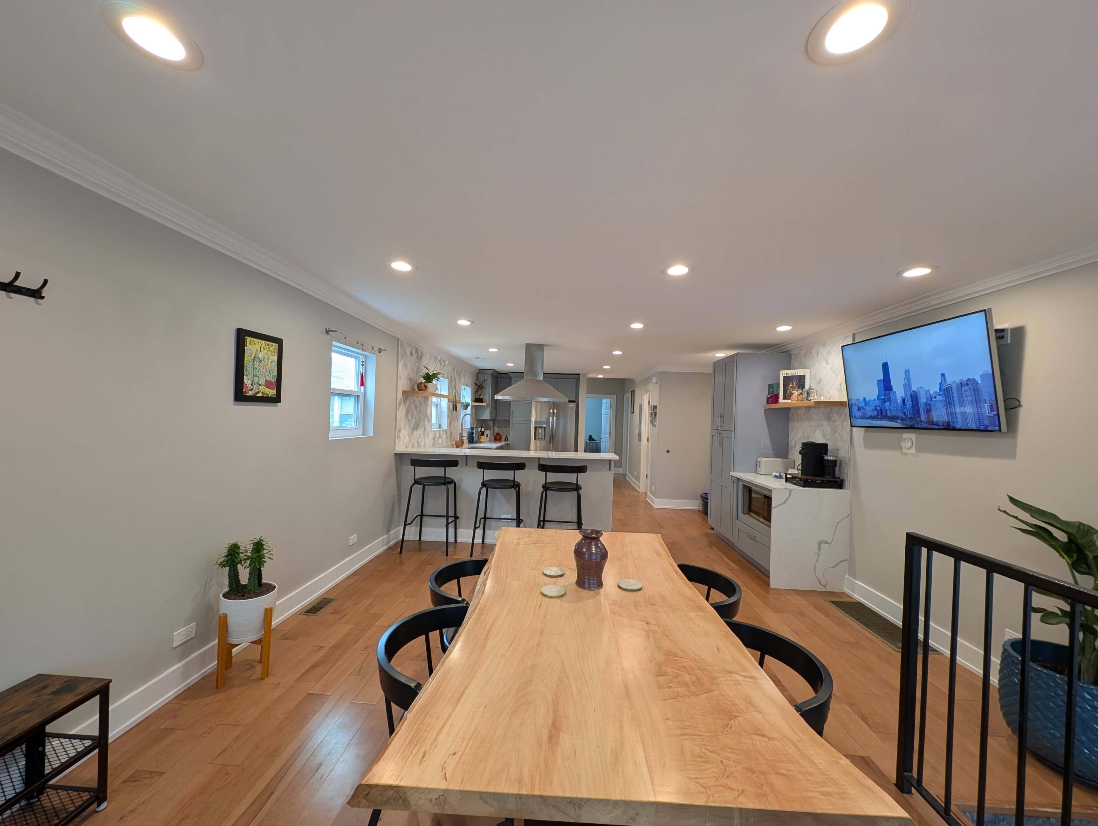 The image shows a modern, open-concept kitchen and dining area with a wooden table, black chairs, and a television mounted on the wall.