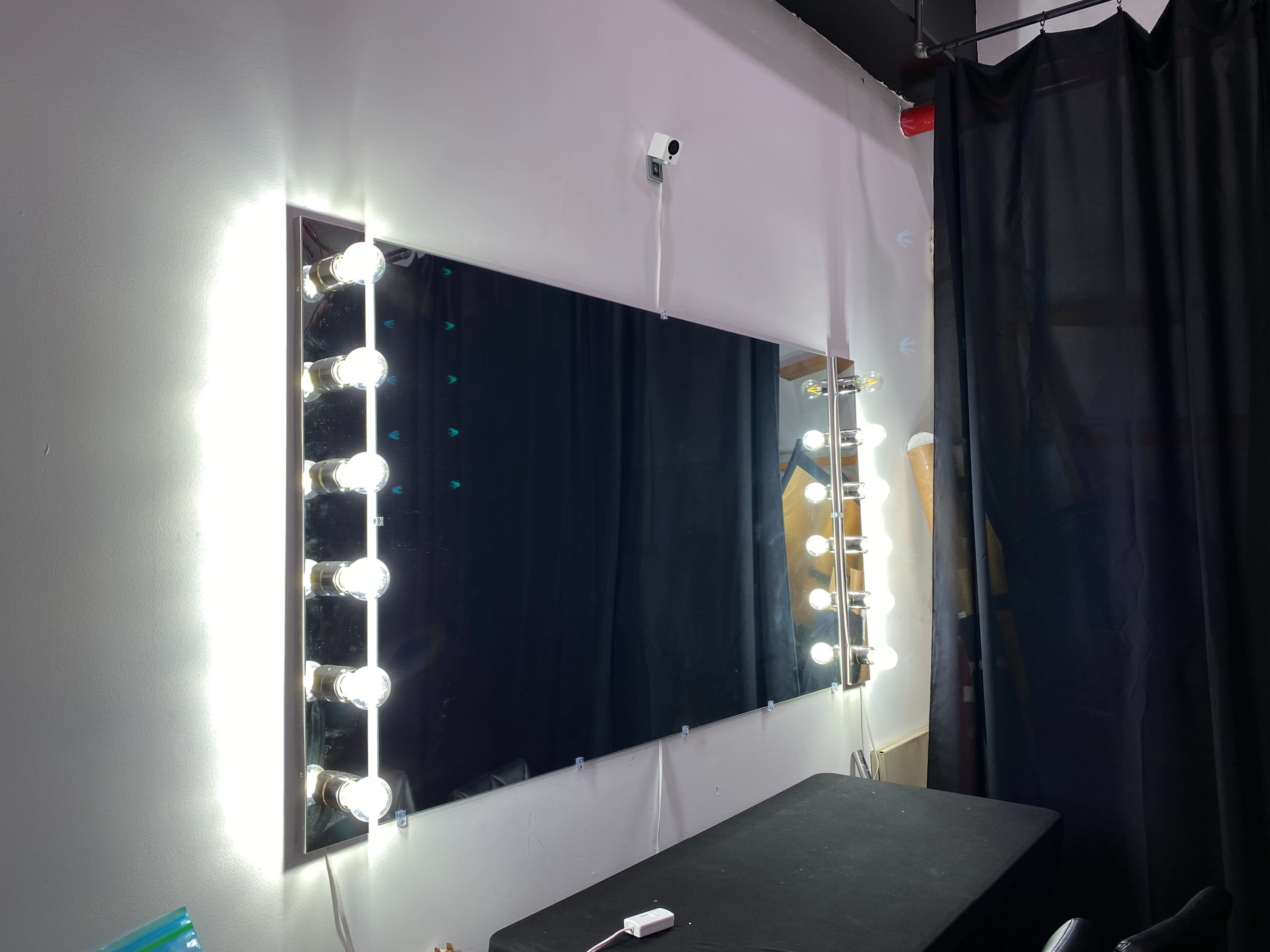 The image shows a wall-mounted mirror surrounded by bright lights, with a black curtain and a table underneath.