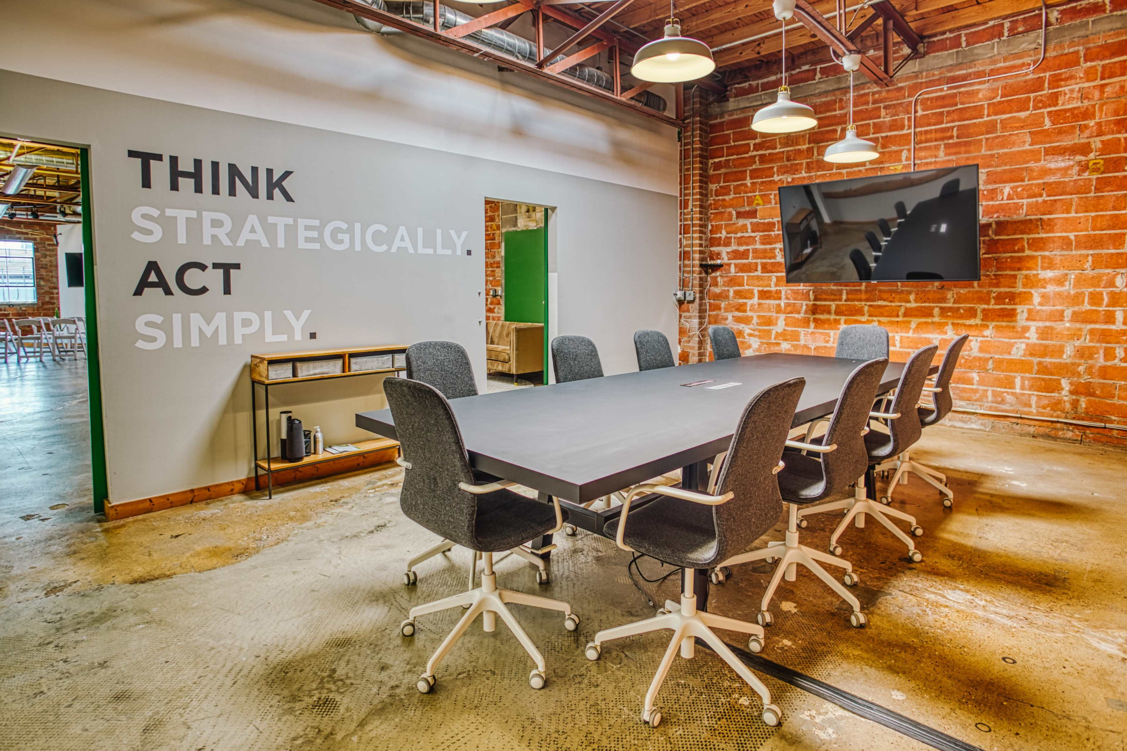 A modern conference room features a long table surrounded by ergonomic chairs, with a large screen mounted on a brick wall and motivational text displayed nearby.
