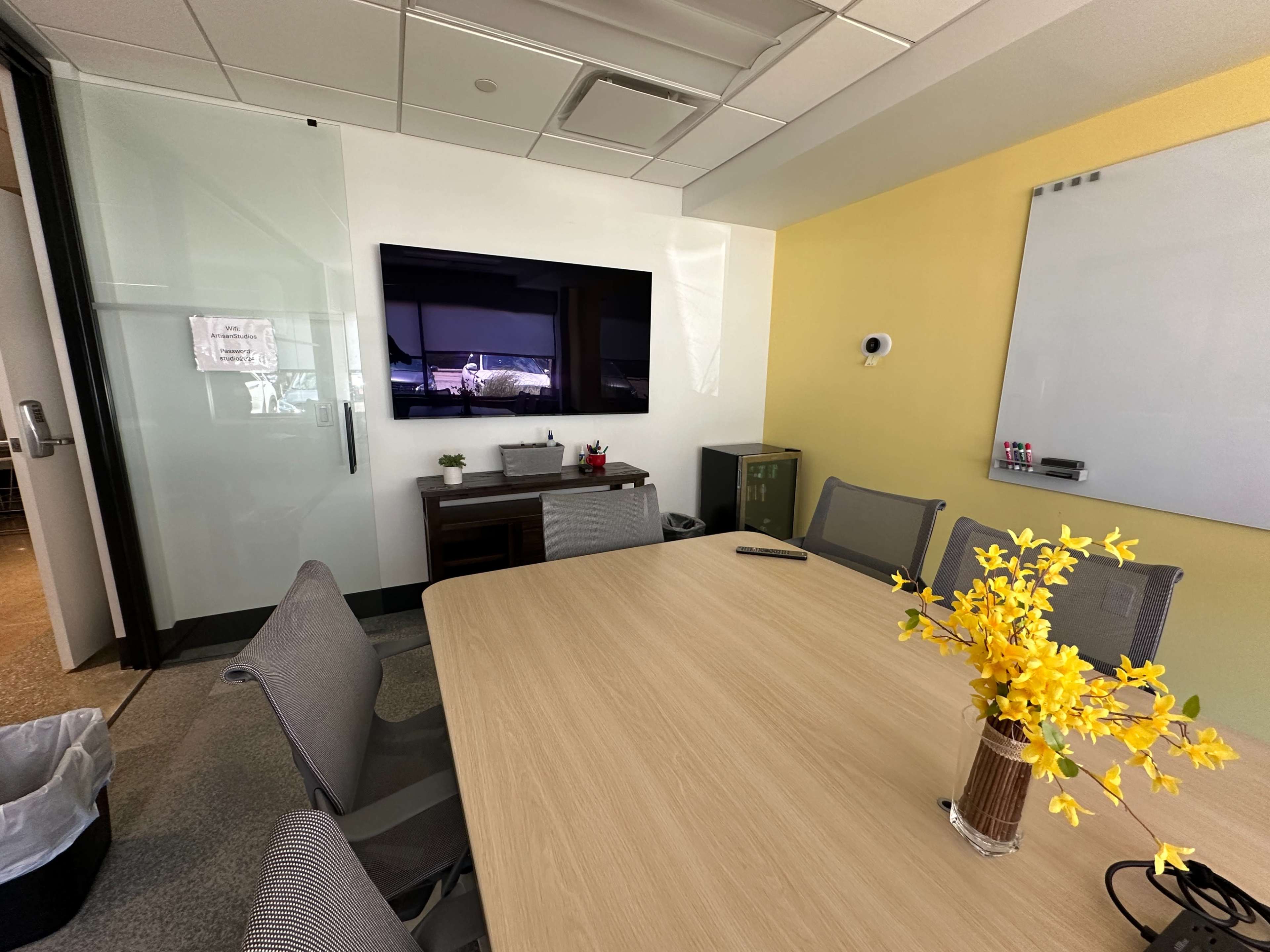 A conference room features a wooden table surrounded by chairs, a wall-mounted screen, and a whiteboard.
