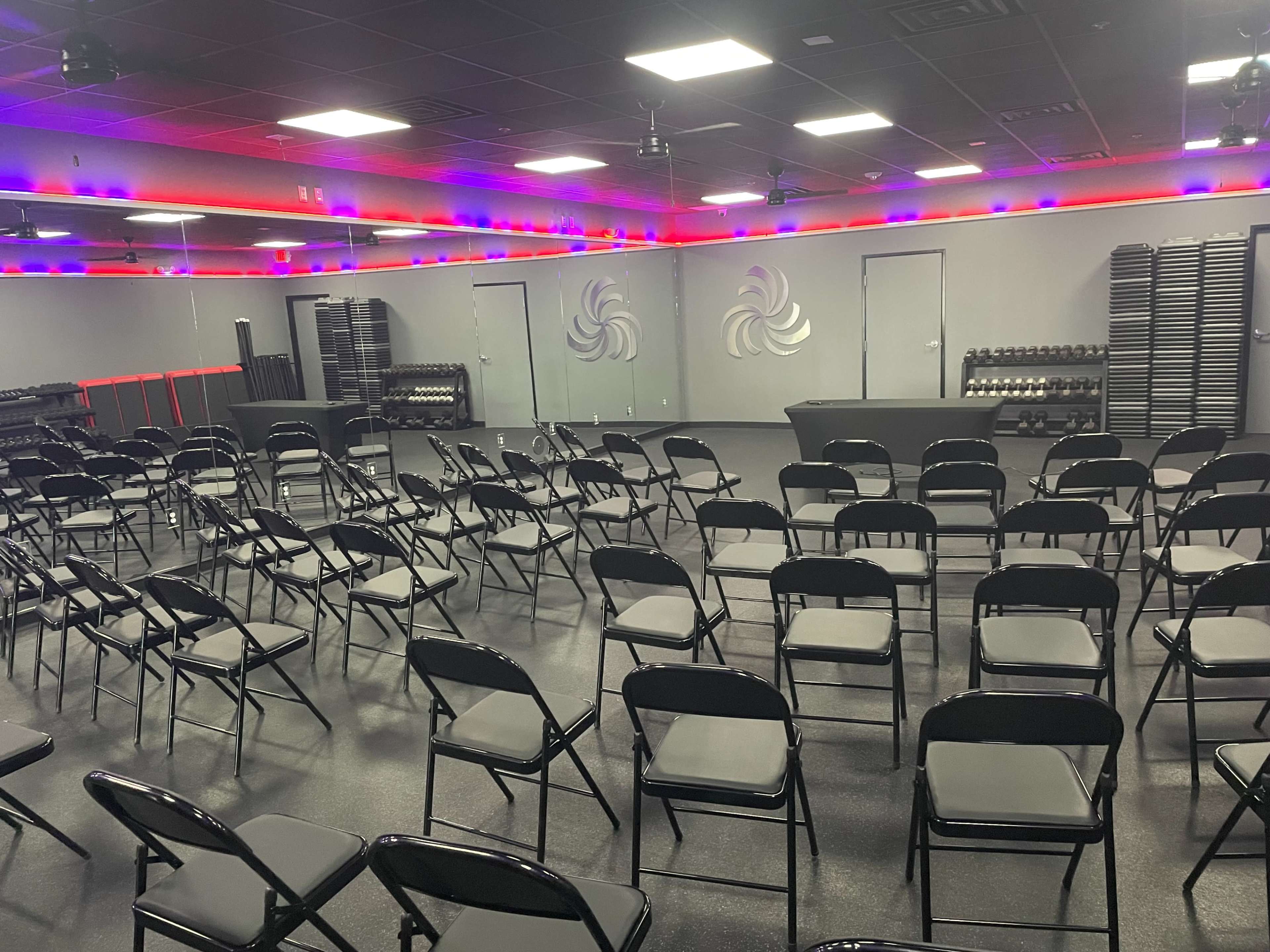 The image shows a spacious room arranged with numerous black folding chairs facing a central stage, with weights and exercise equipment neatly stacked against the walls.