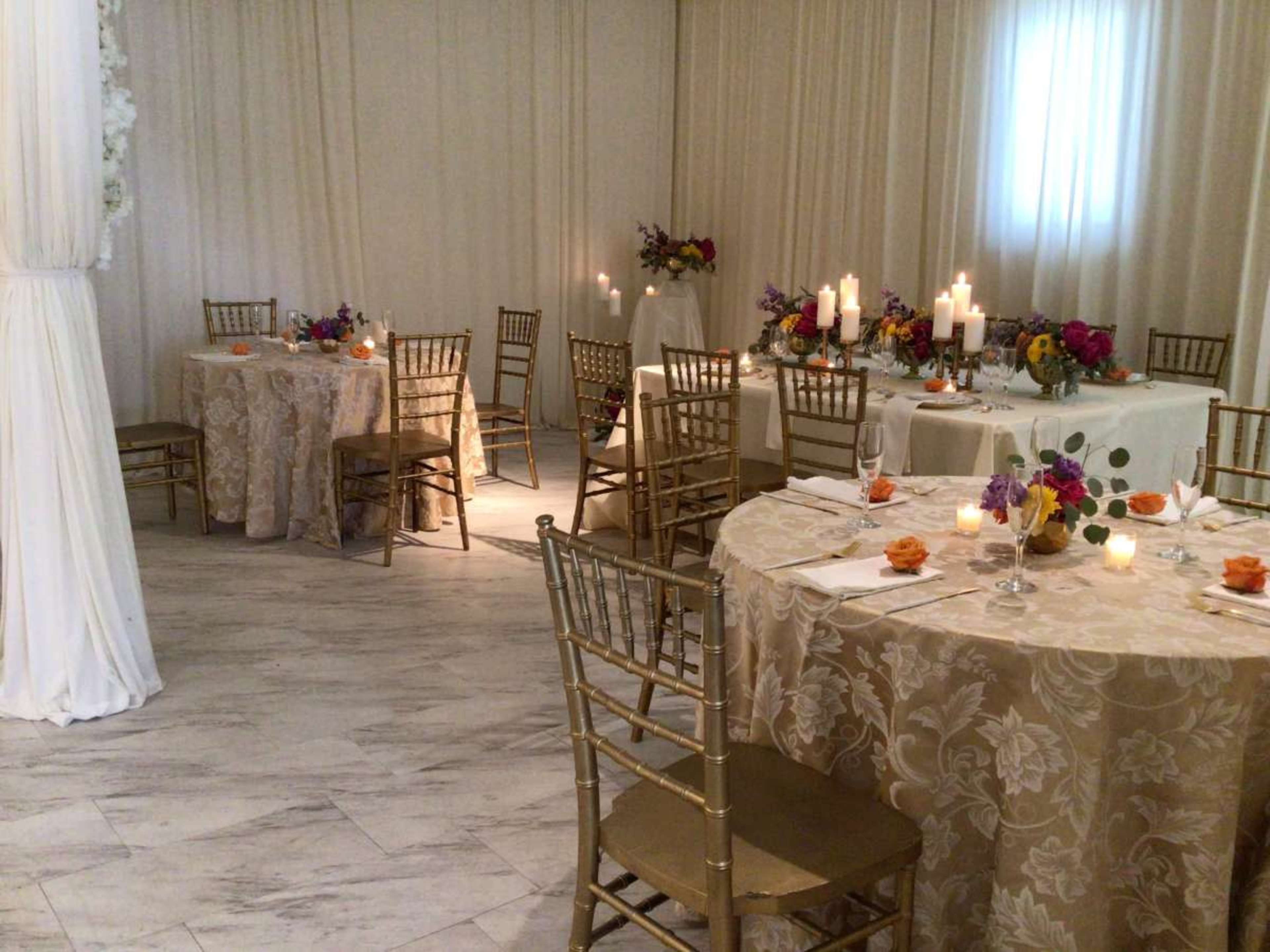 The image shows an elegantly set dining area with tables adorned in patterned tablecloths, candles, and floral centerpieces, surrounded by gold chairs.