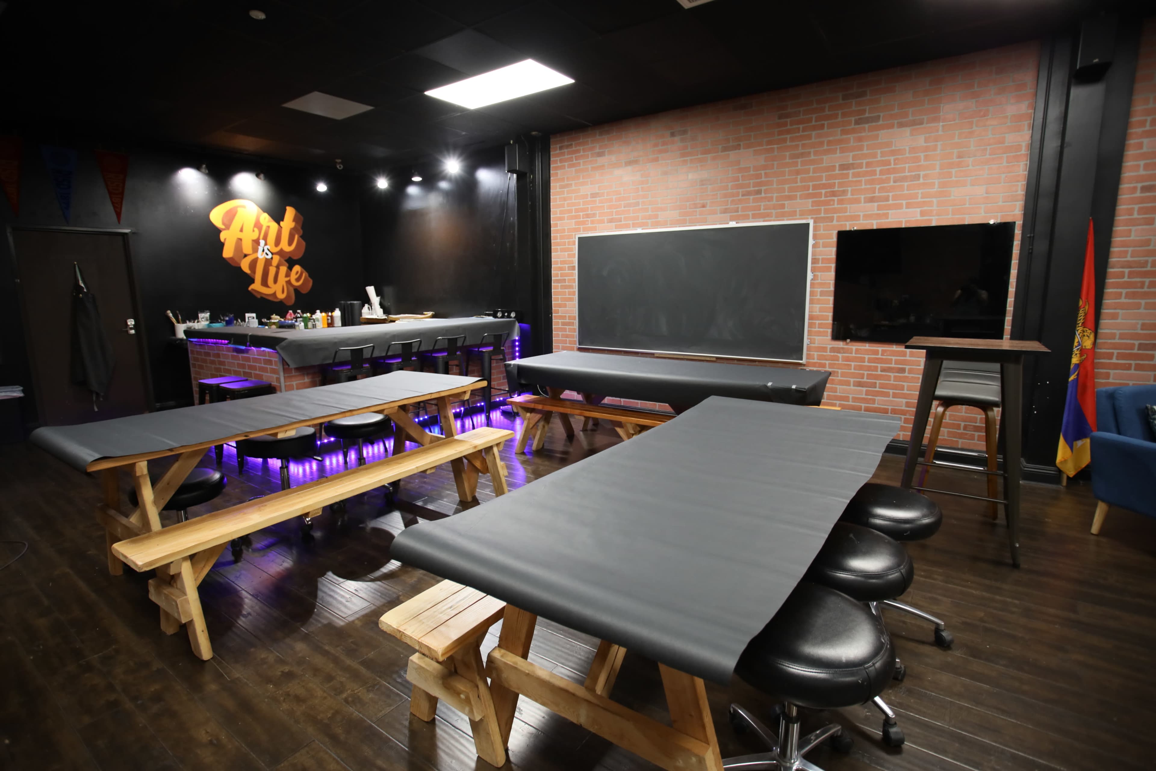 An art studio with several wooden tables covered in black paper, a chalkboard, and a bar area illuminated with purple lighting.