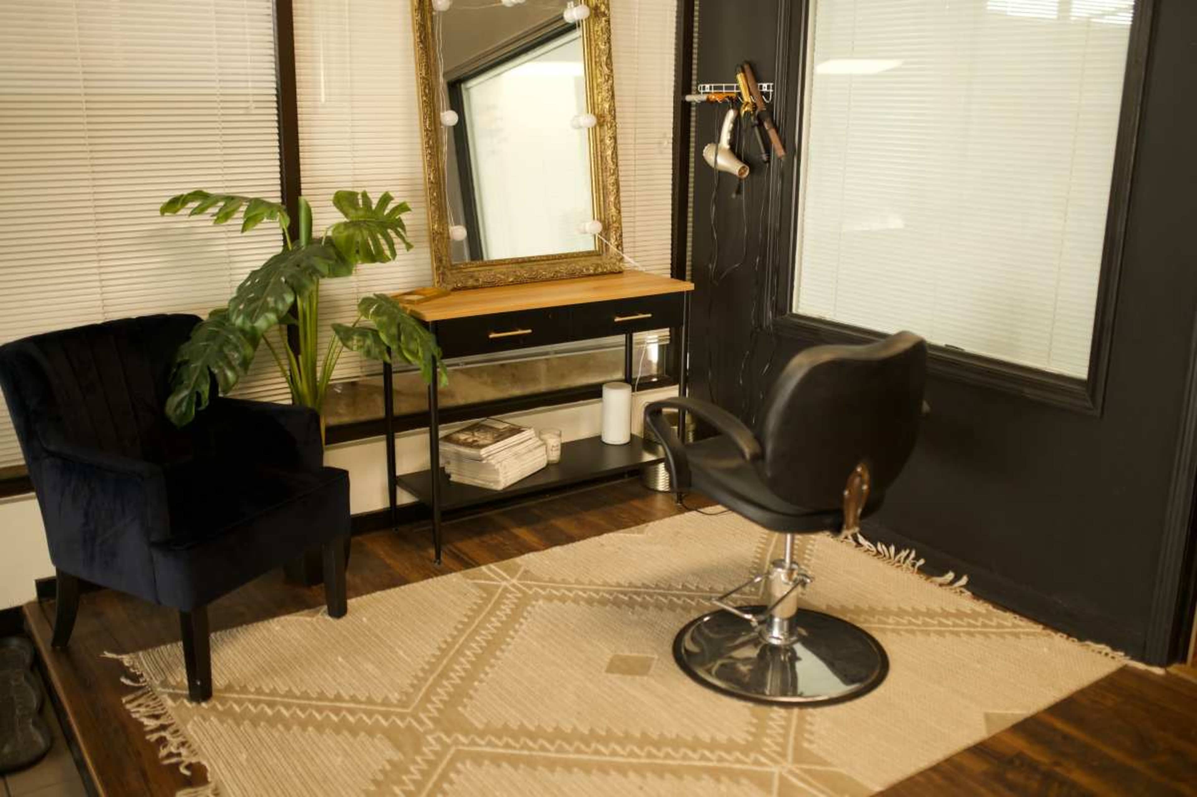 The image shows a small hair salon setup featuring a black styling chair, a gold-framed mirror, a plant, and a storage console with a few magazines on a patterned rug.