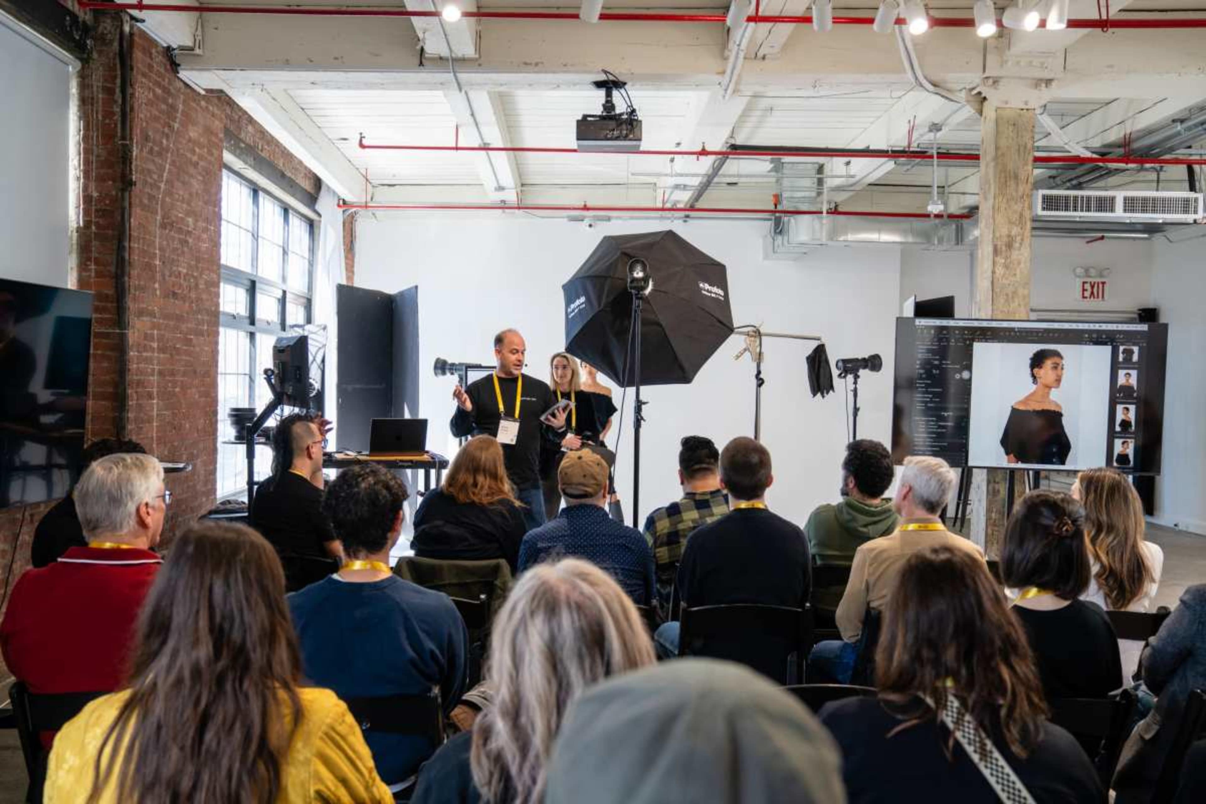 A group of people is seated in a photography studio, watching a presentation by two speakers in front of a backdrop and studio lighting.