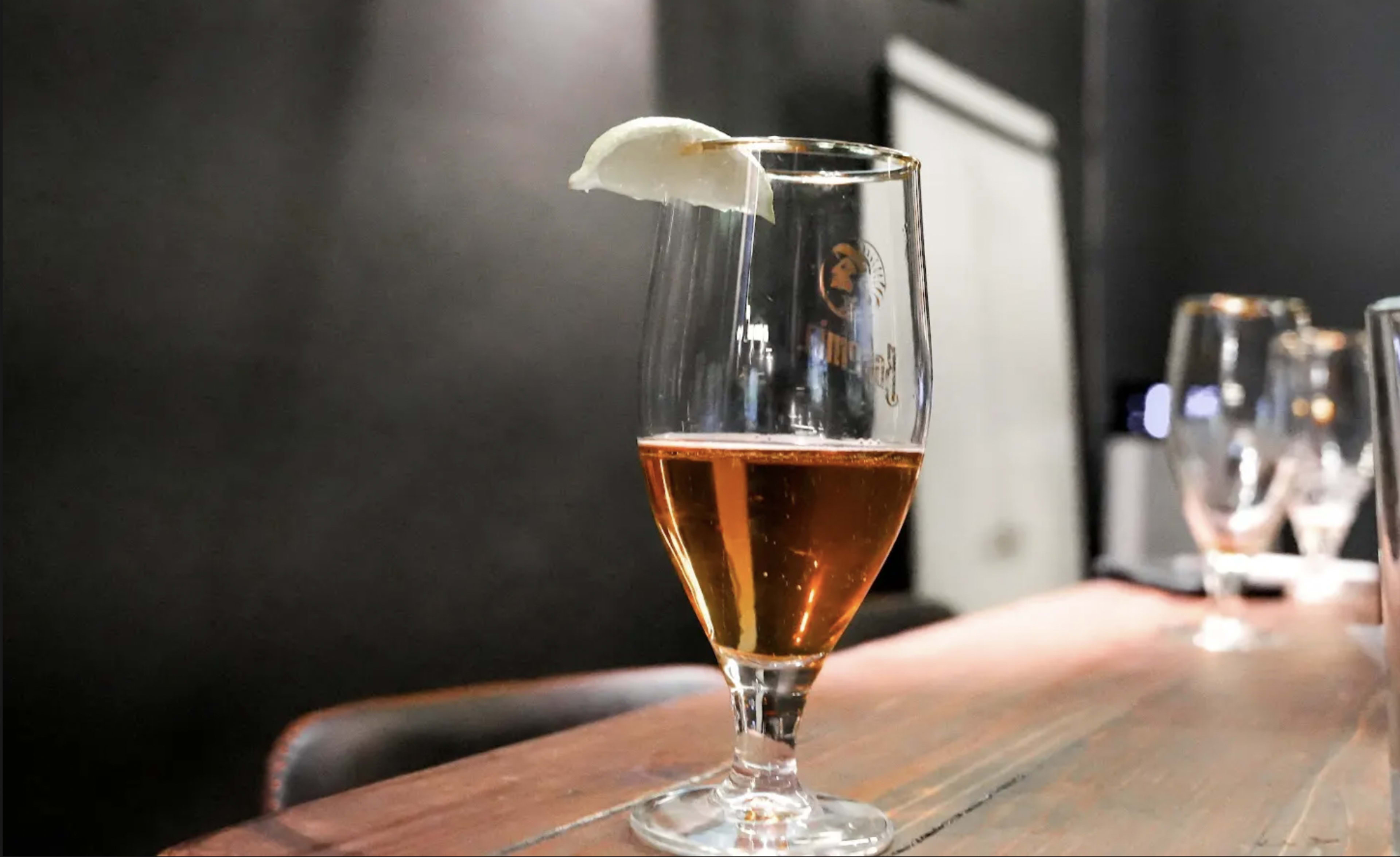 A glass of amber beer garnished with a slice of lemon sits on a wooden table in a dimly lit environment.