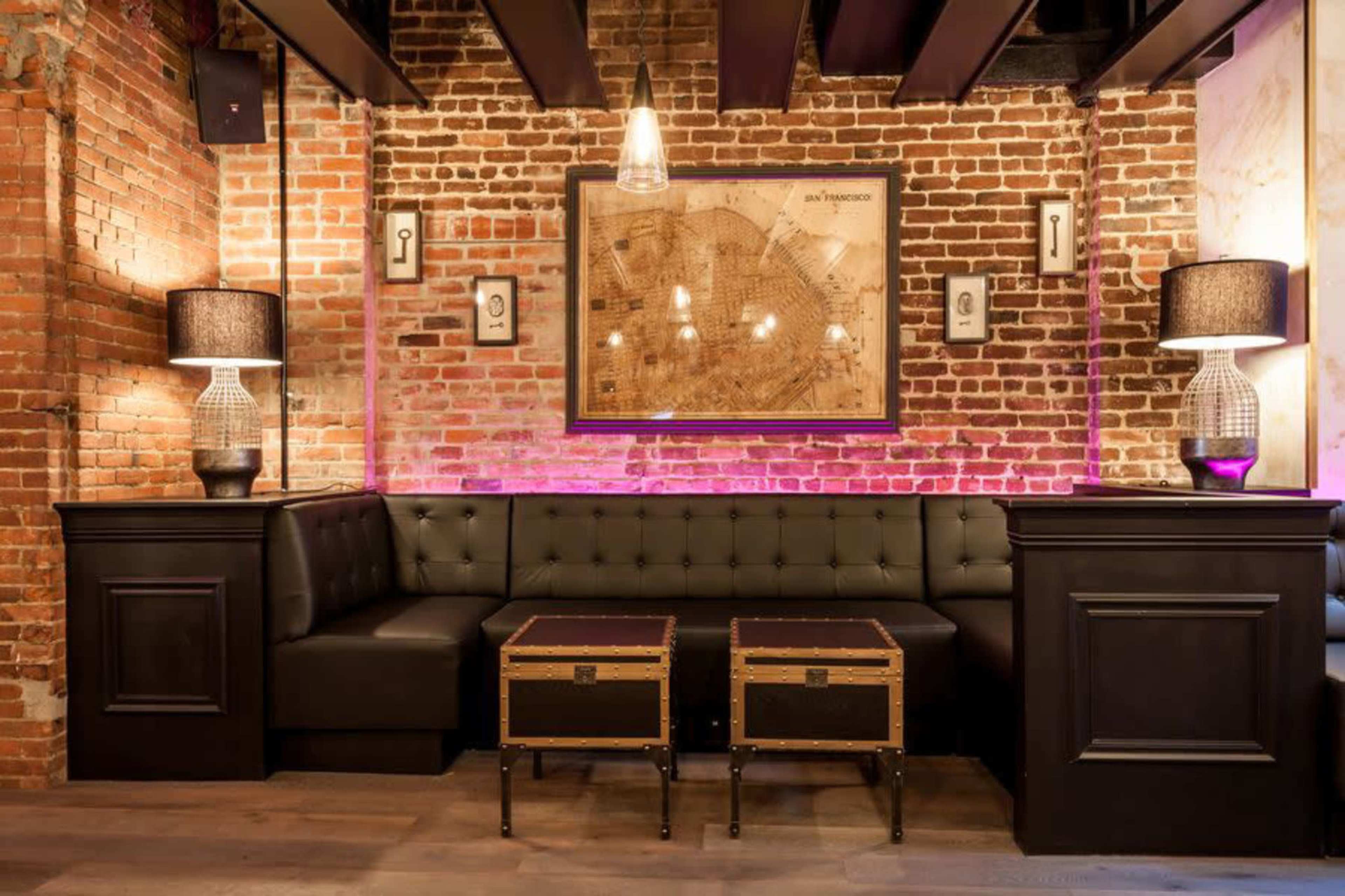 The image shows a cozy seating area in a restaurant with a tufted black leather couch, two wooden trunk-style tables, and exposed brick walls illuminated by soft lighting.