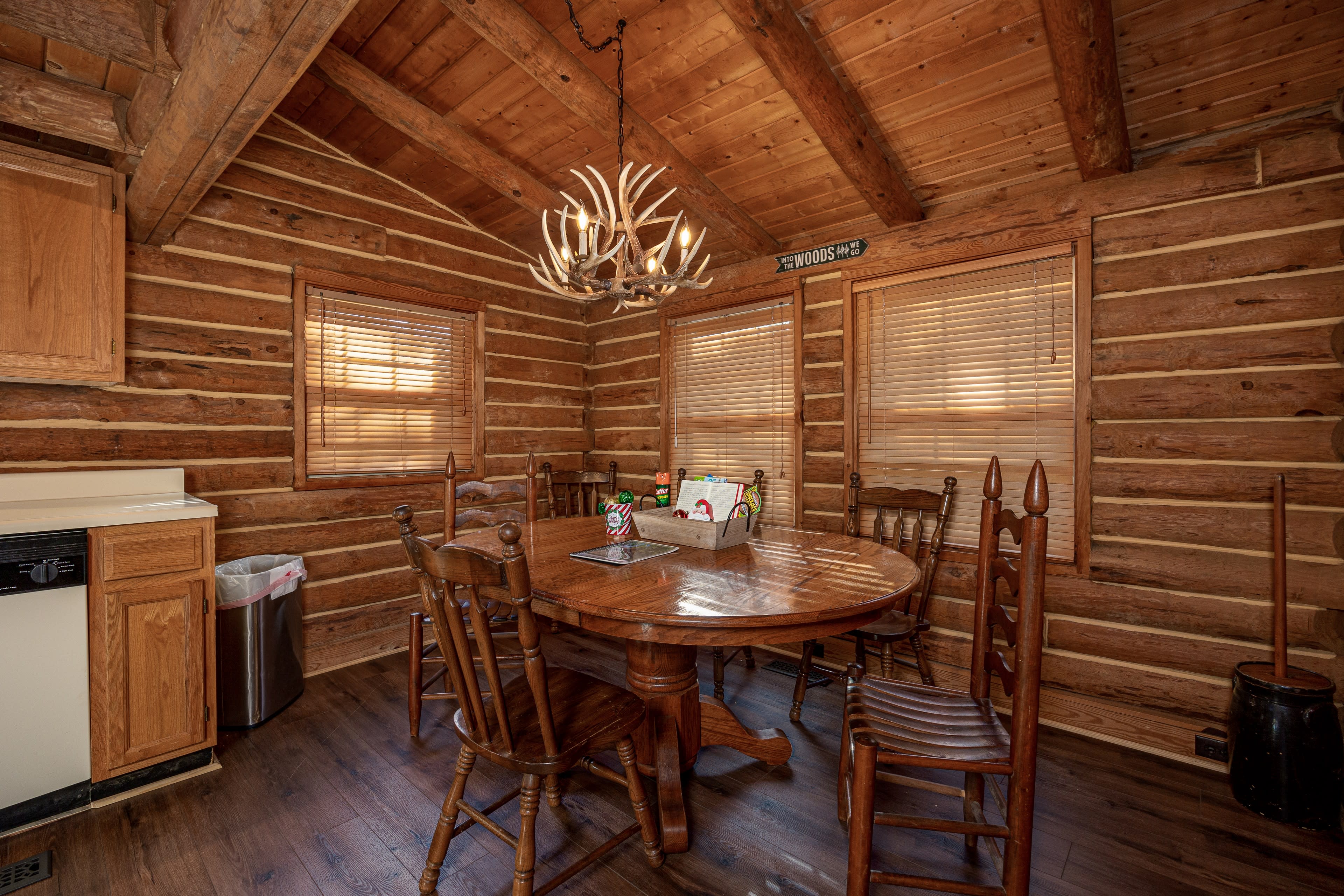 The image shows a rustic dining area with a round wooden table surrounded by several wooden chairs under a chandelier made of antlers, set against log cabin walls.